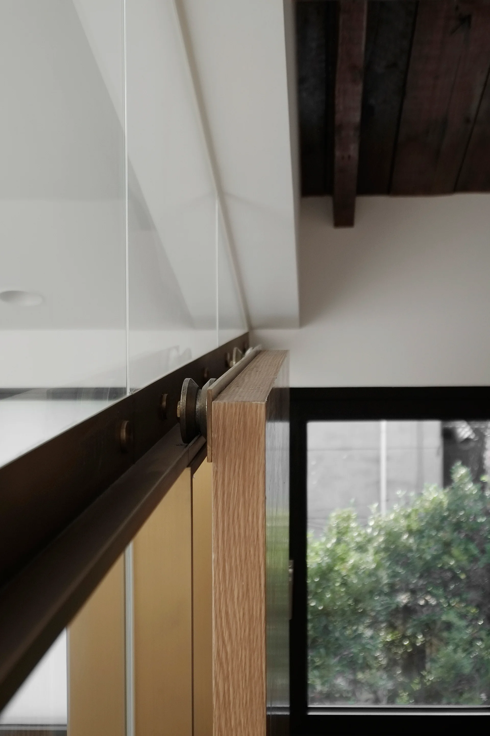 A close-up of a light oak sliding barn door hanging from a dark metal track roller mechanism in a room with a white wall.