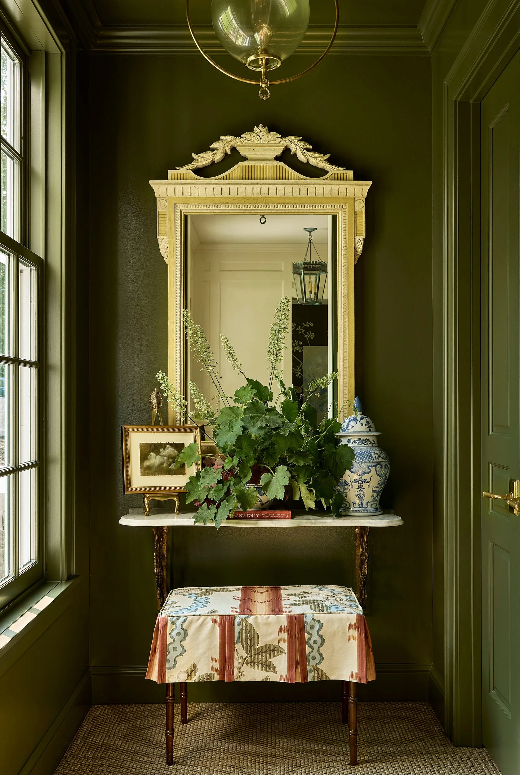An entryway vestibule painted in deep olive green features a marble console table, an ornate gold mirror, and a patterned upholstered stool.