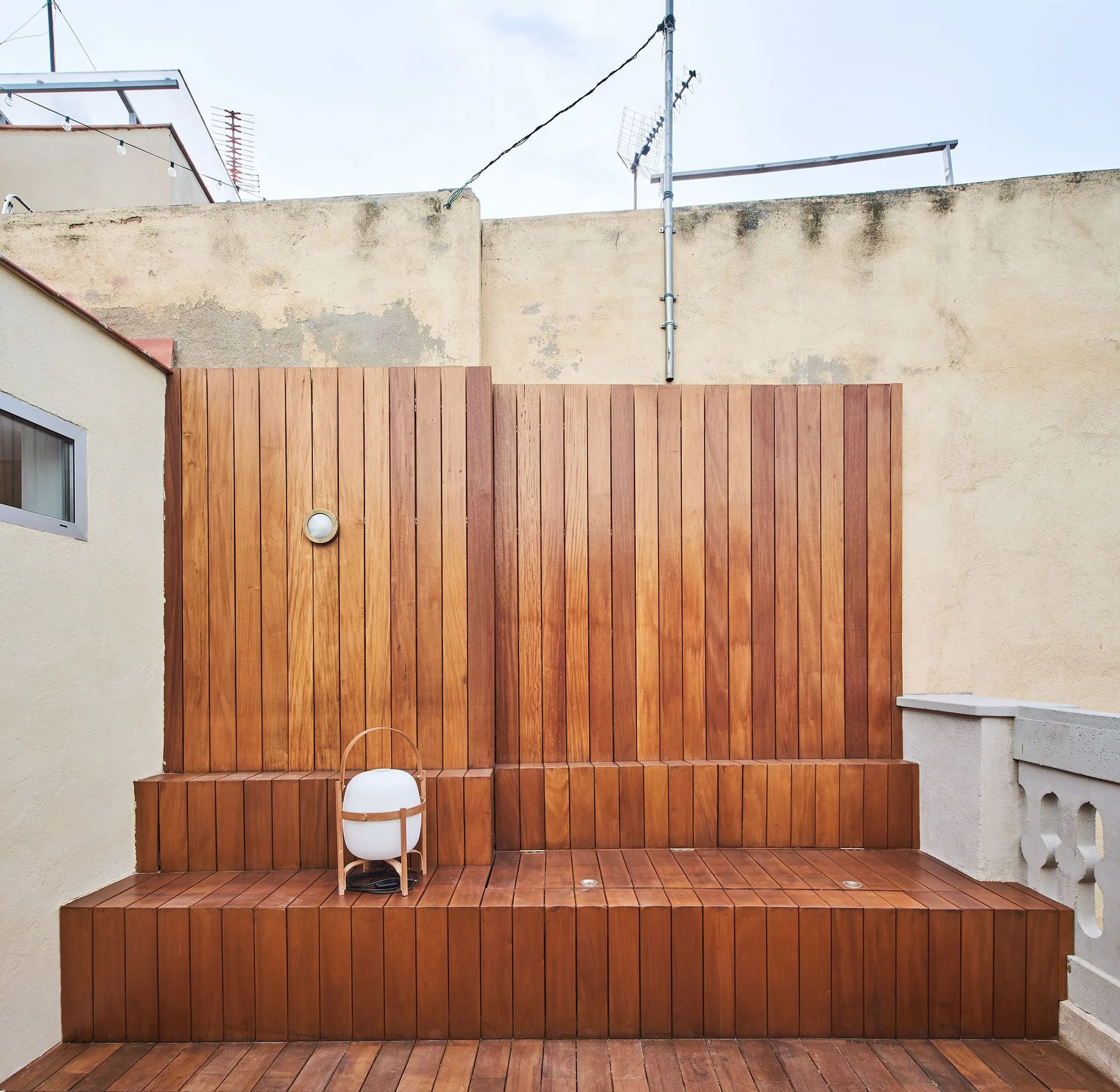A rooftop terrace featuring a tiered wooden deck with matching vertical wood cladding, a portable lantern, and integrated floor lighting.