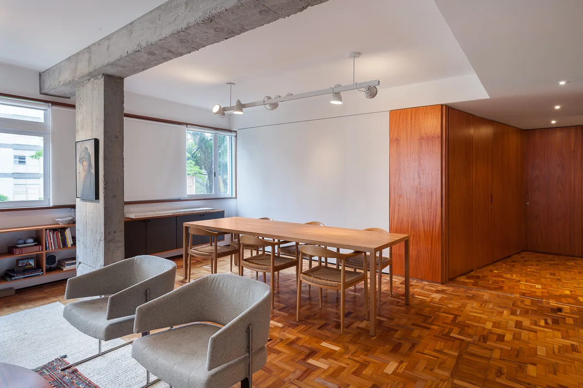 A bright dining area features a wooden table and chairs on parquet flooring, next to an exposed concrete column and a wood-paneled wall.