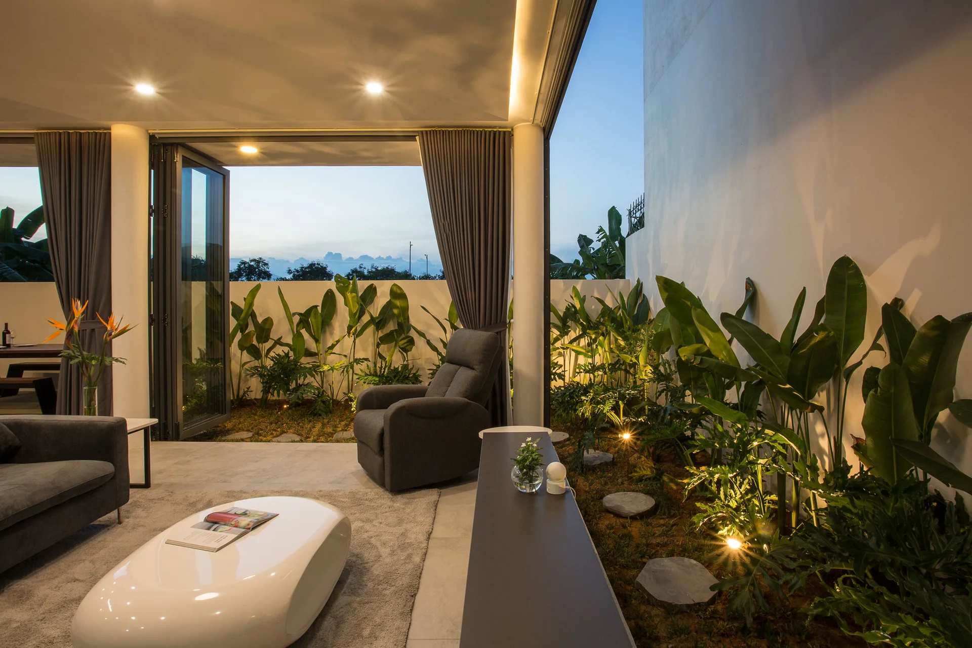 A grey sofa and white oval coffee table in a modern living room that opens to an outdoor courtyard with green plants and landscape lighting.