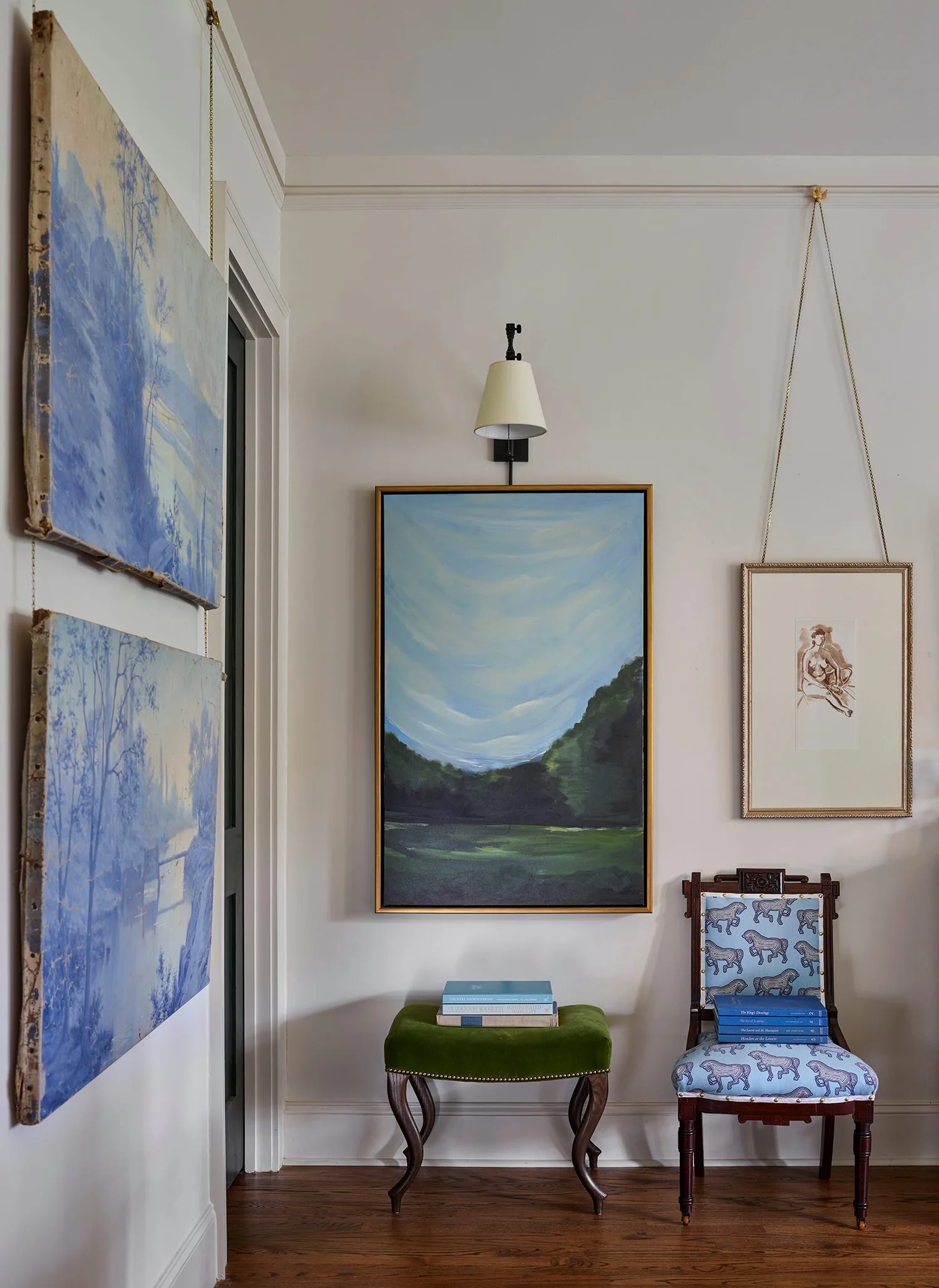 A curated gallery wall features landscape paintings hung on chains, a green velvet stool, and a blue patterned chair on a hardwood floor.