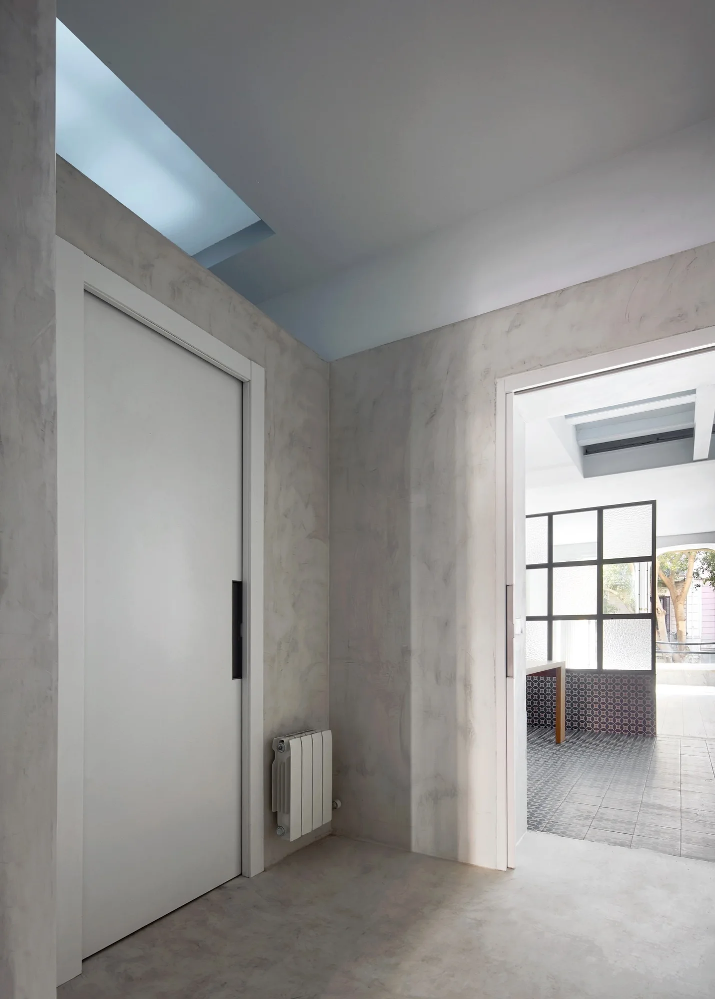A minimalist hallway featuring textured grey plaster walls, a white sliding door, a ceiling skylight, and a view into a room with patterned tile floors.