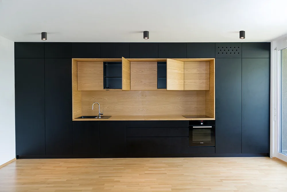 A modern kitchen wall featuring matte black handleless cabinets and a central light wood recessed niche containing a sink, cooktop, and matching upper cabinets.