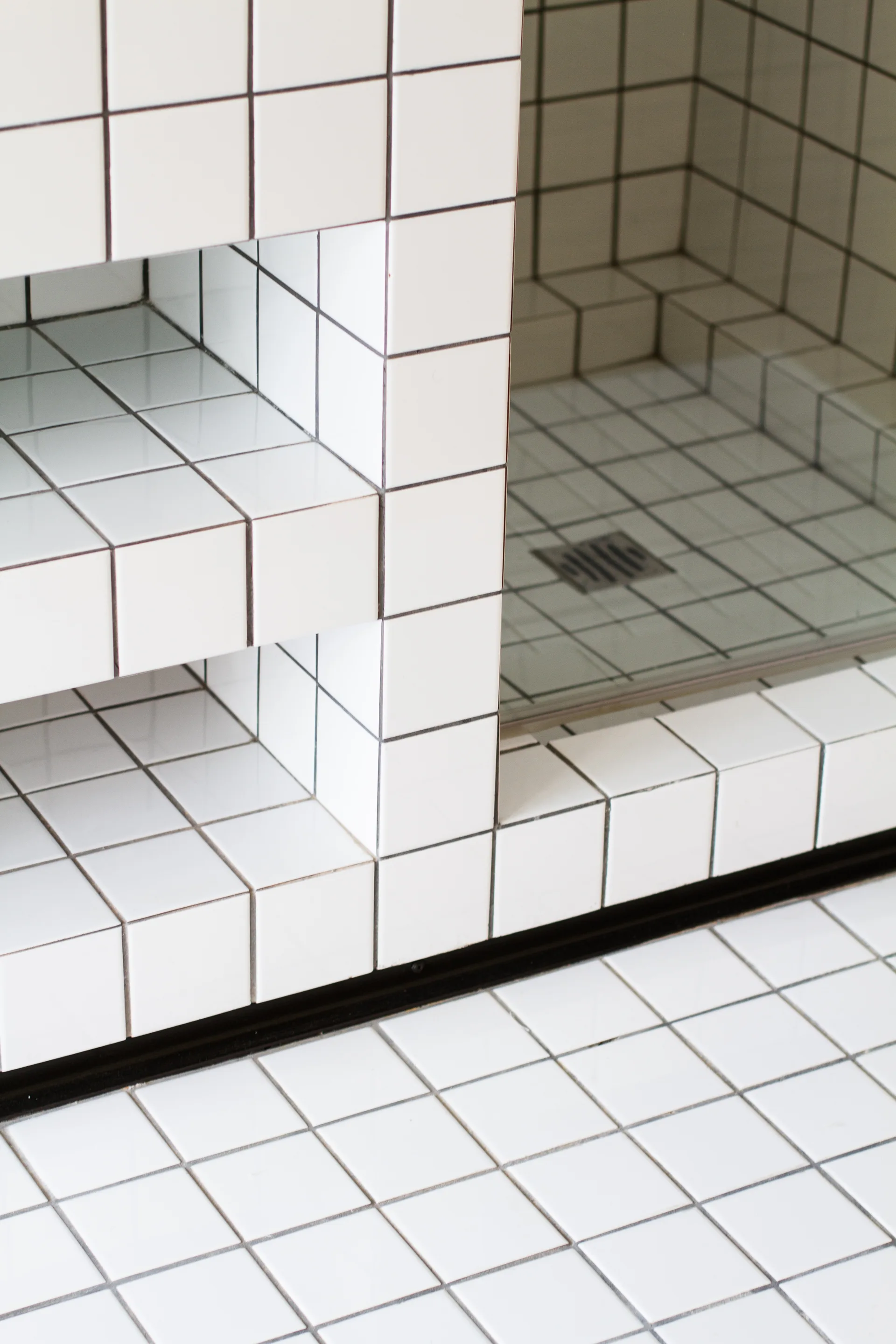 A close-up of a bathroom shower showing white square grid tiles with black grout on the floor, wall, and built-in storage unit.