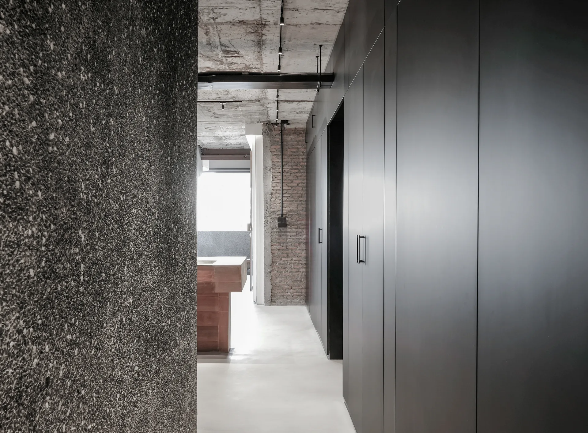A modern industrial hallway features full-height charcoal gray cabinetry on one side, an exposed concrete ceiling, and a raw brick accent wall.