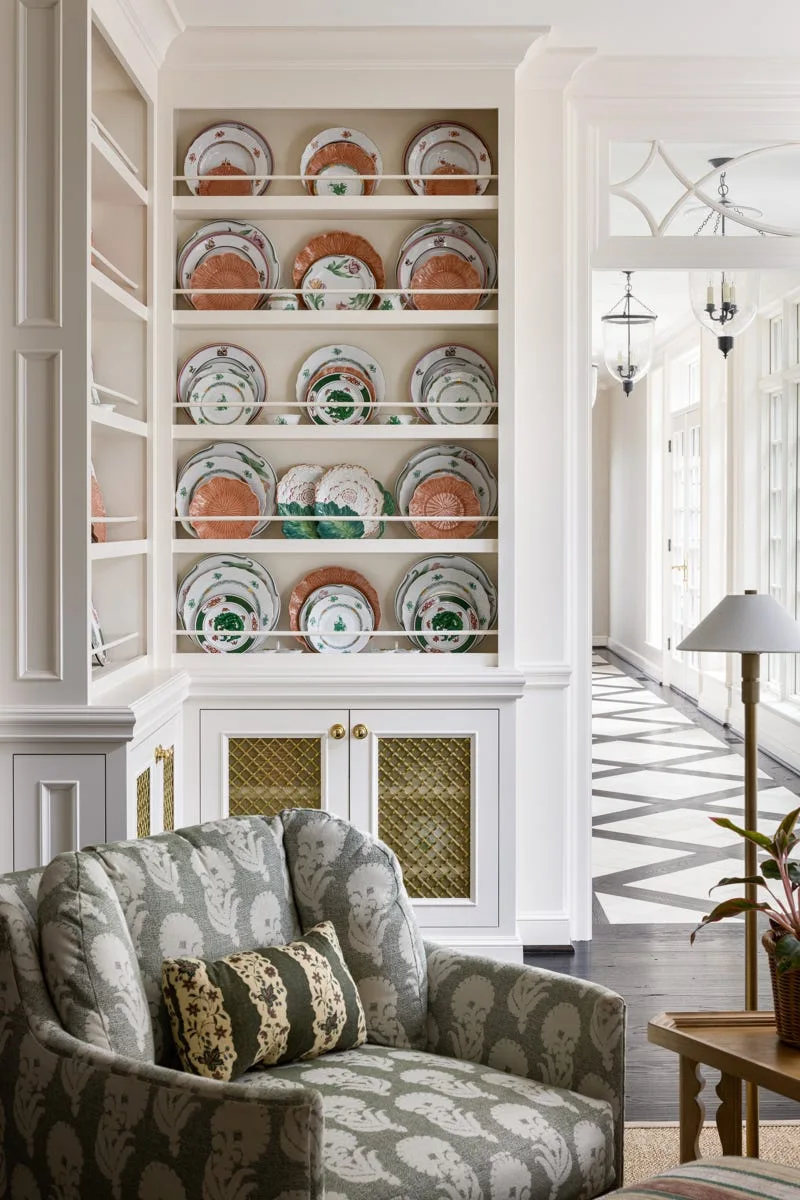 A patterned green armchair sits near a white built-in cabinet featuring shelves lined with green and terracotta decorative plates.