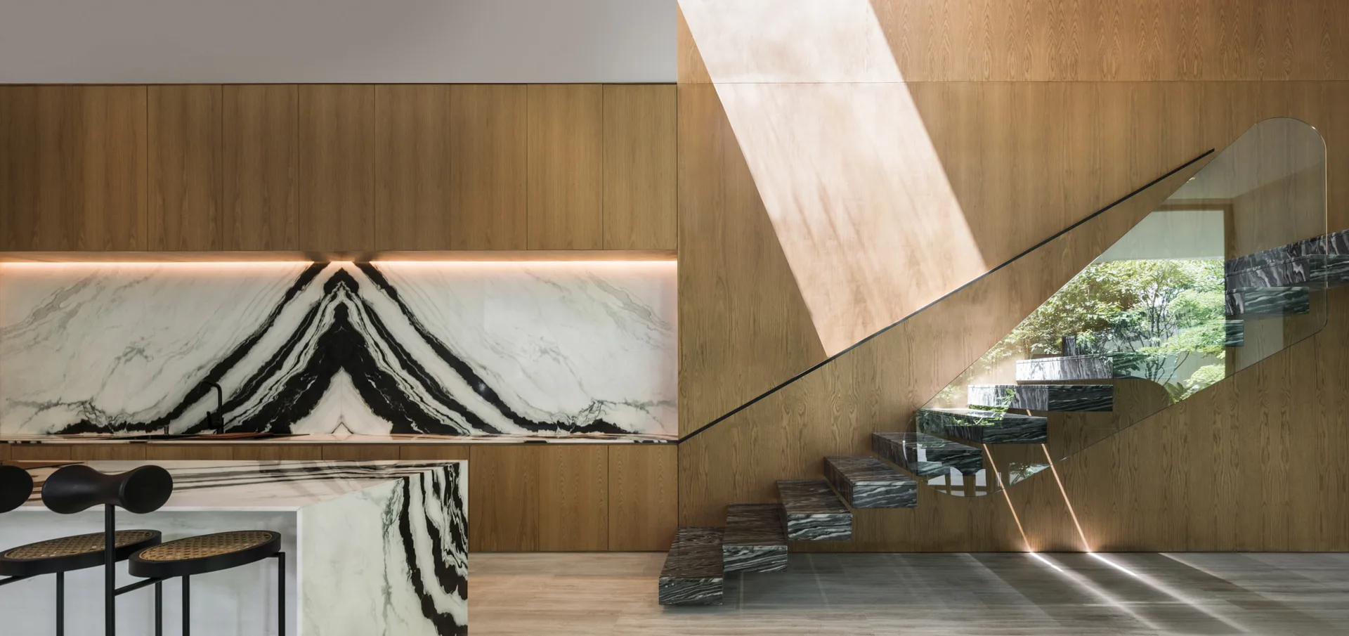 A modern kitchen features warm wood cabinetry, a book-matched marble backsplash, a marble island, and floating marble stairs with a glass railing.