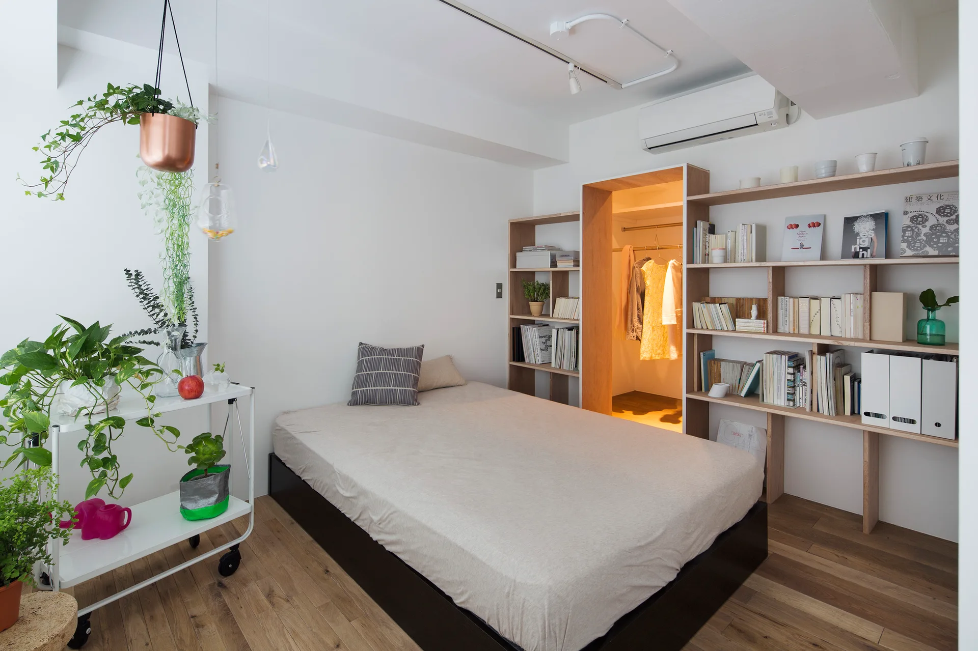 A modern minimalist bedroom features a platform bed flanked by light wood open bookcases built around a glowing walk-in closet entryway.