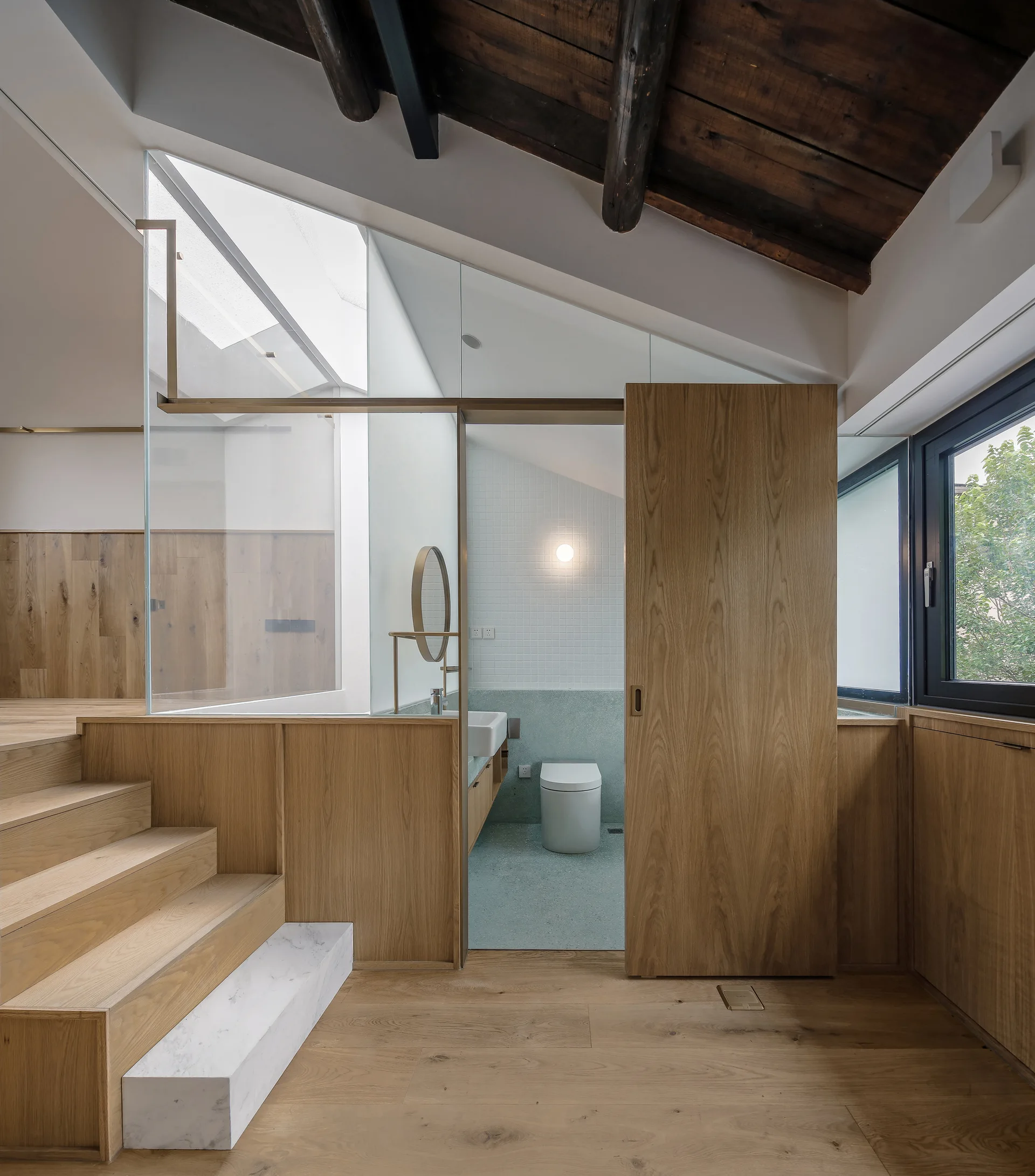 A modern loft space with wooden stairs, a marble accent step, and a glass-enclosed bathroom featuring a wooden sliding door under exposed timber beams.