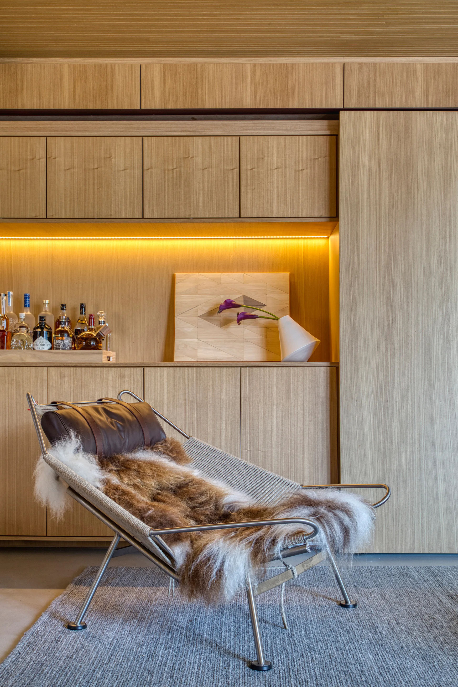A modern rope and metal lounge chair with a fur throw stands before light wood bar cabinetry featuring integrated warm LED lighting.