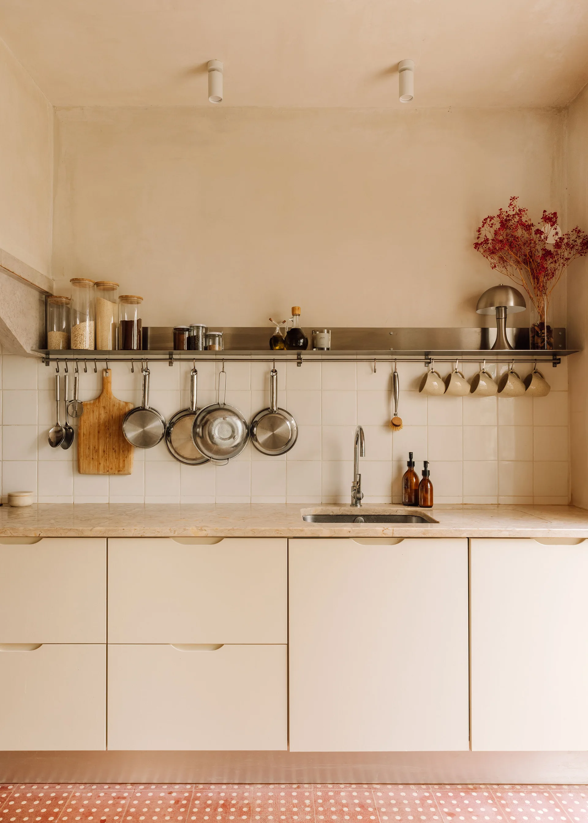 A minimalist kitchen features flat cream cabinets, a beige stone countertop, and a white tiled backsplash with a stainless steel storage rail and shelf.