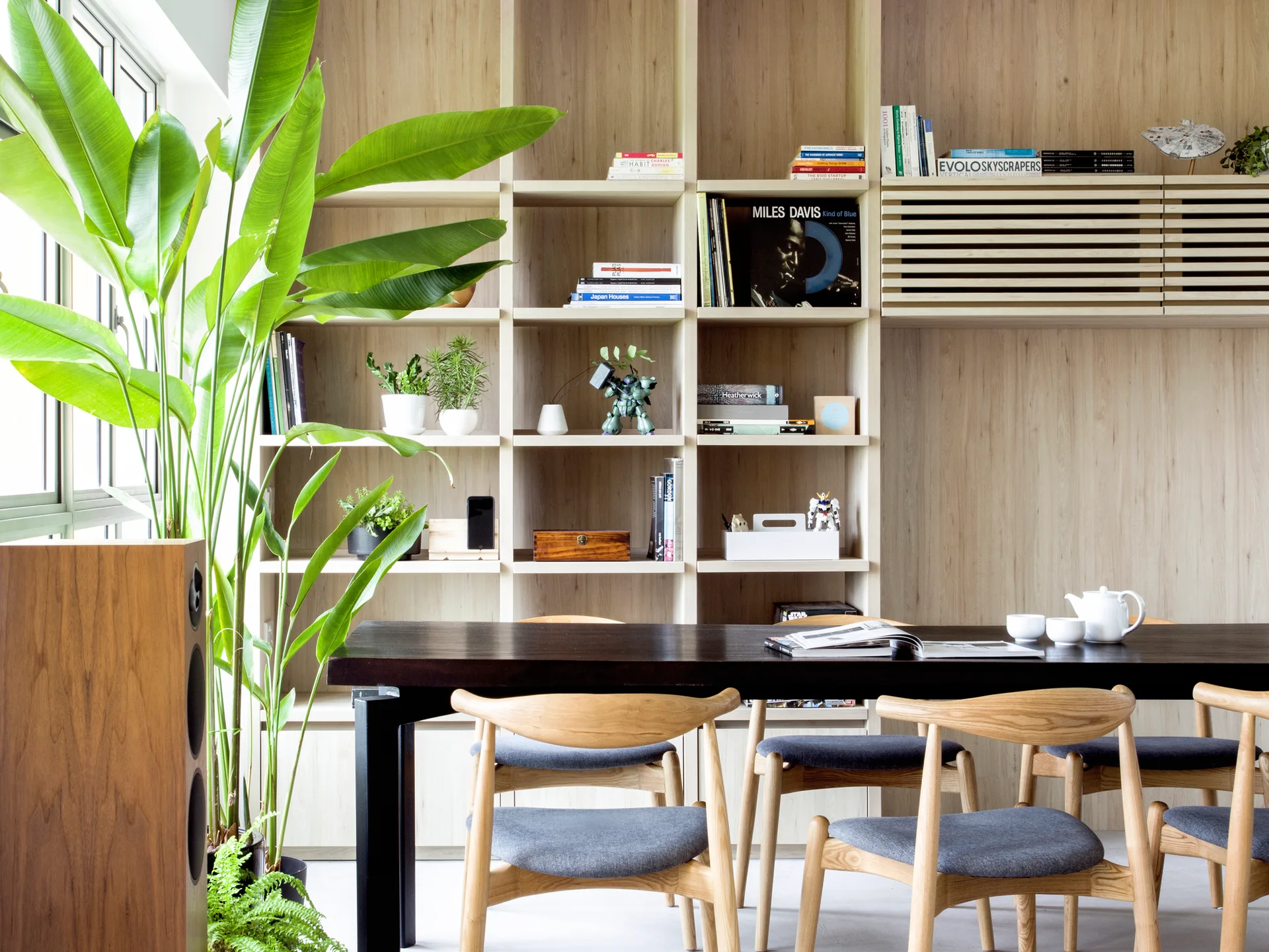A dark wood dining table and light oak chairs sit in front of a floor-to-ceiling light wooden shelving unit filled with books and plants.