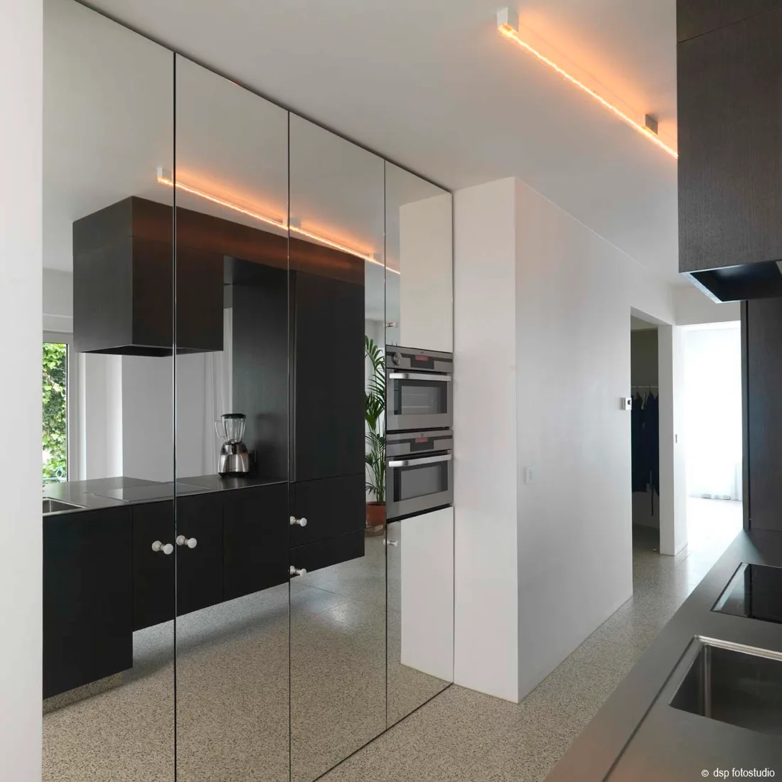 A modern kitchen features tall mirrored cabinets reflecting dark countertops and a potted plant, alongside white walls and a speckled terrazzo floor.