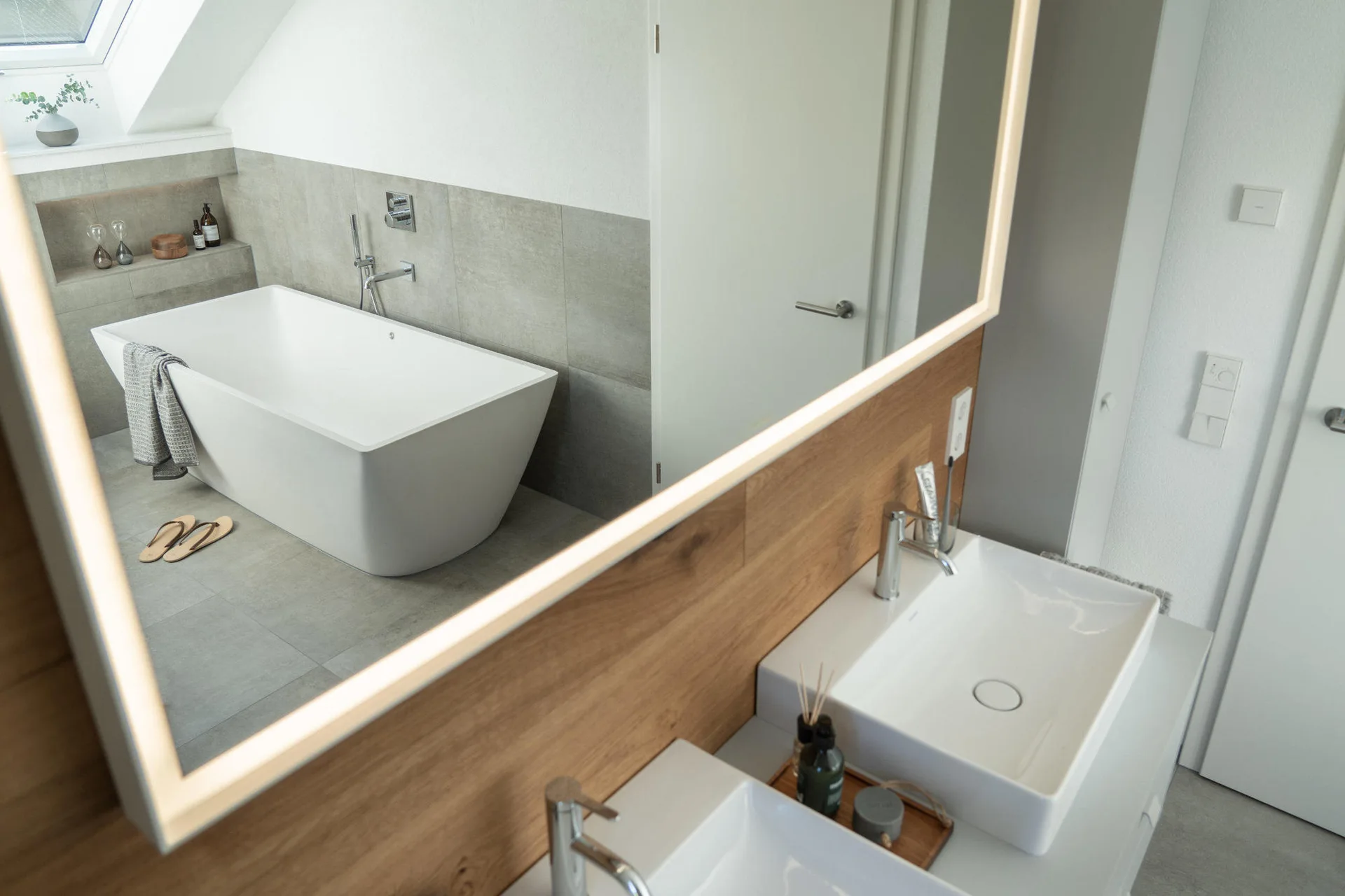 A modern bathroom mirror with integrated LED lighting reflects a freestanding white bathtub under a sloped ceiling with a skylight.
