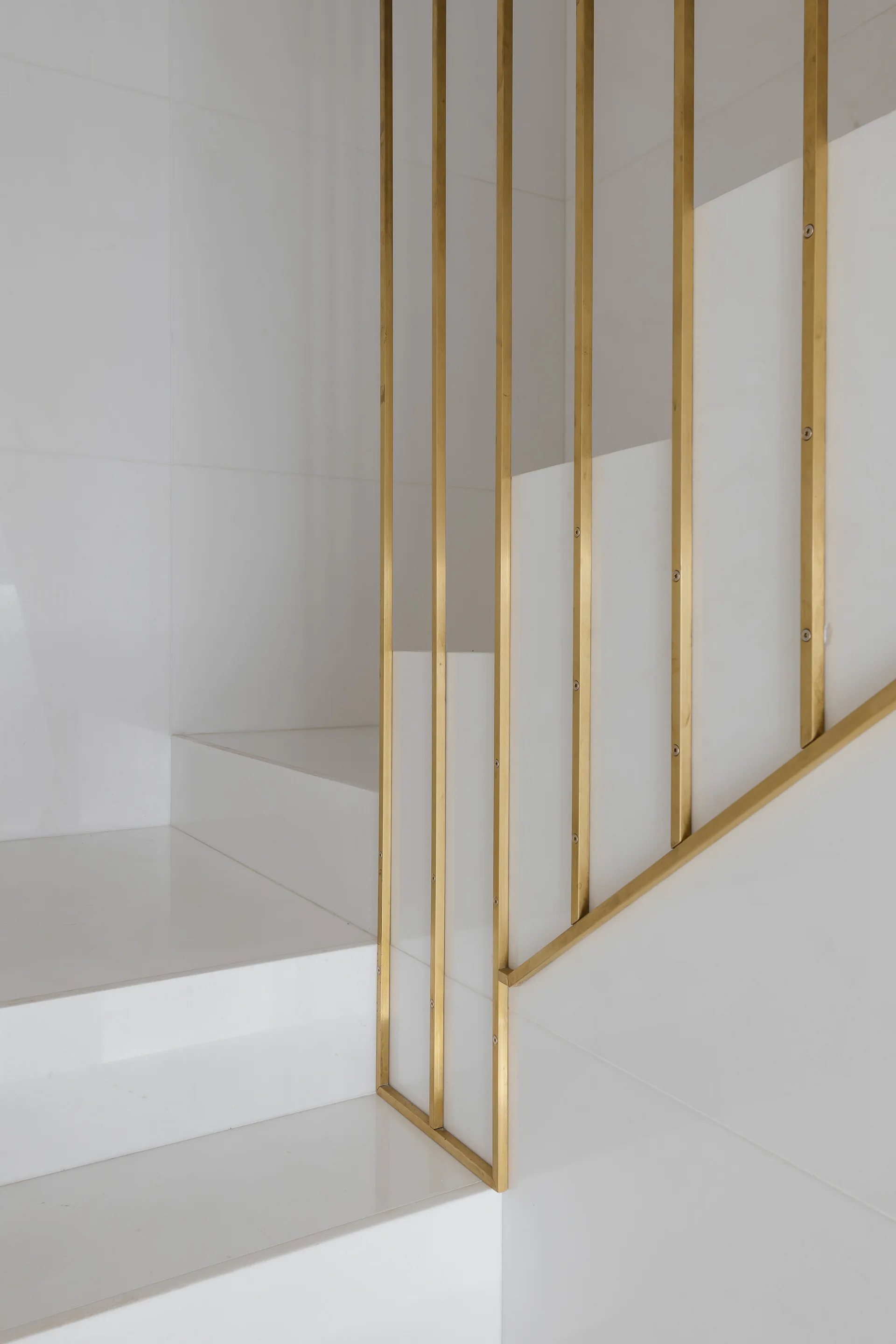 Sleek white staircase steps are flanked by a modern vertical railing made of thin brass metal bars.