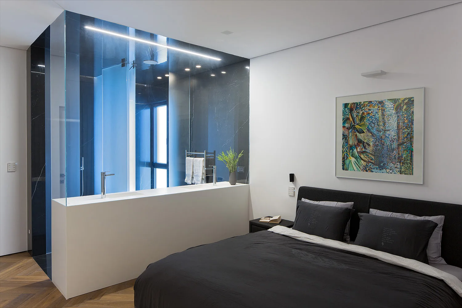 A contemporary bedroom features a dark bed and an integrated open bathroom vanity with a glass partition and herringbone wood floors.
