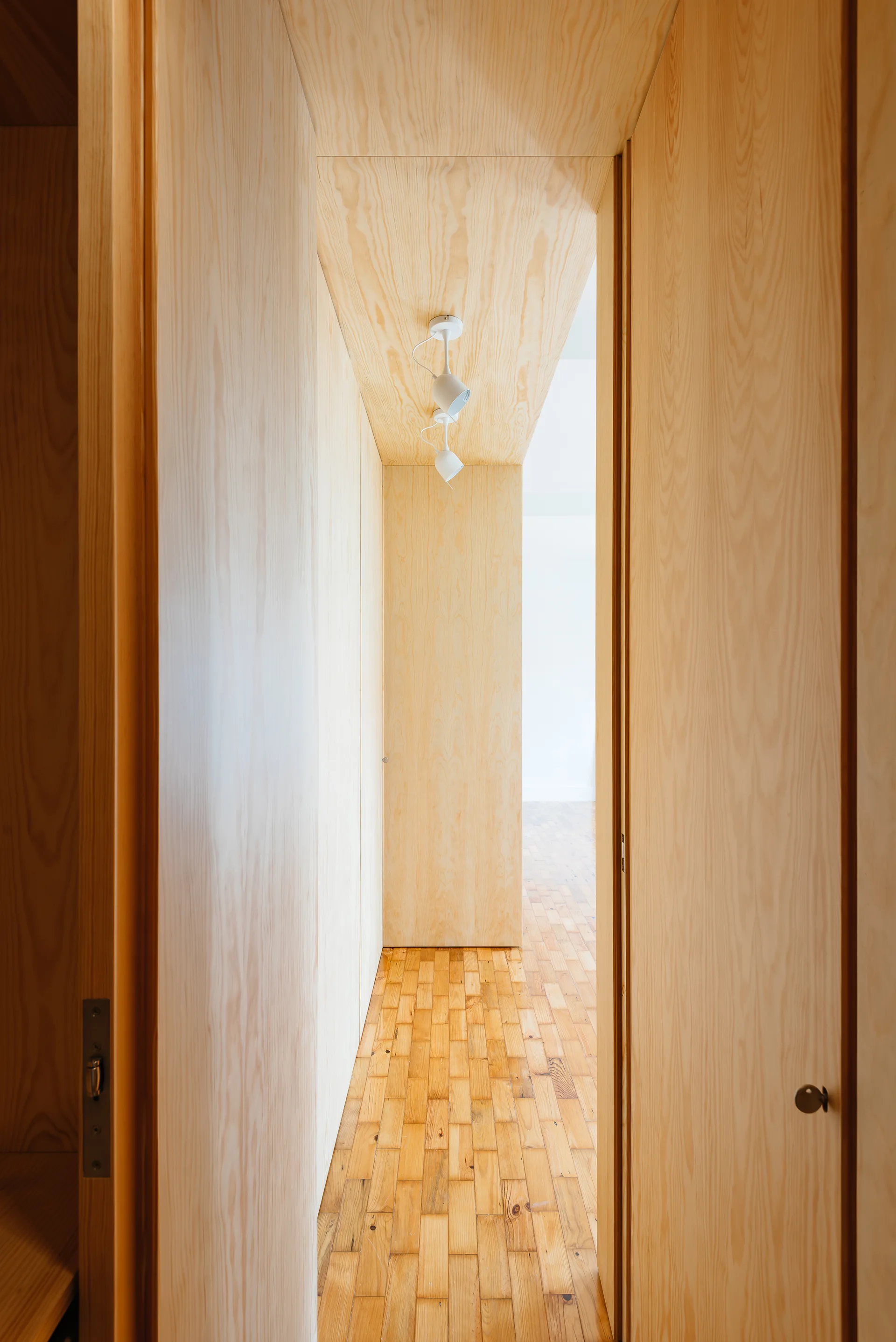 A narrow hallway featuring light wood-veneer walls and ceiling, a parquet wood floor, and a white ceiling-mounted spotlight.