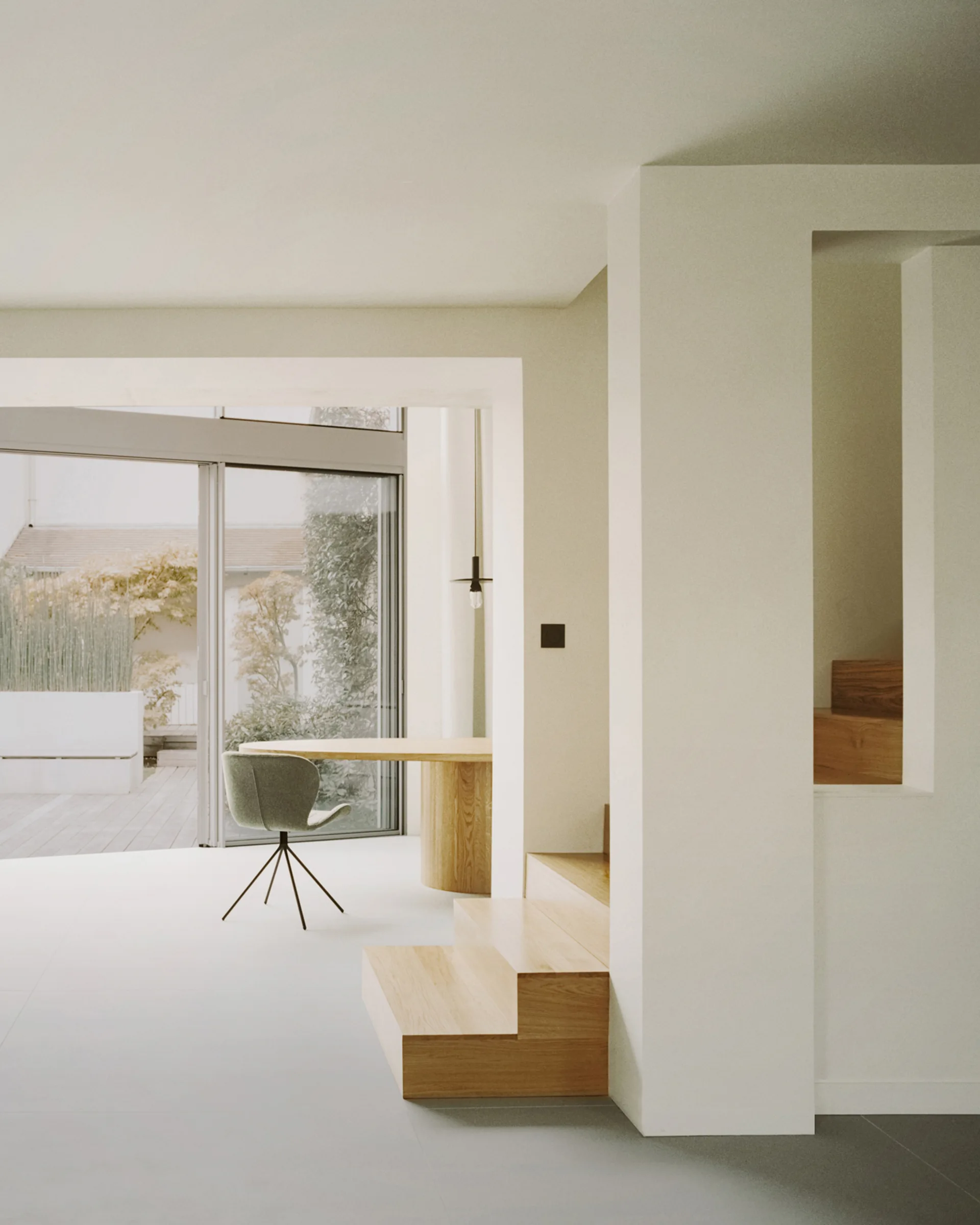 A minimalist interior featuring a light wood staircase, a round wooden table with a sage green chair, and glass sliding doors opening to a patio.