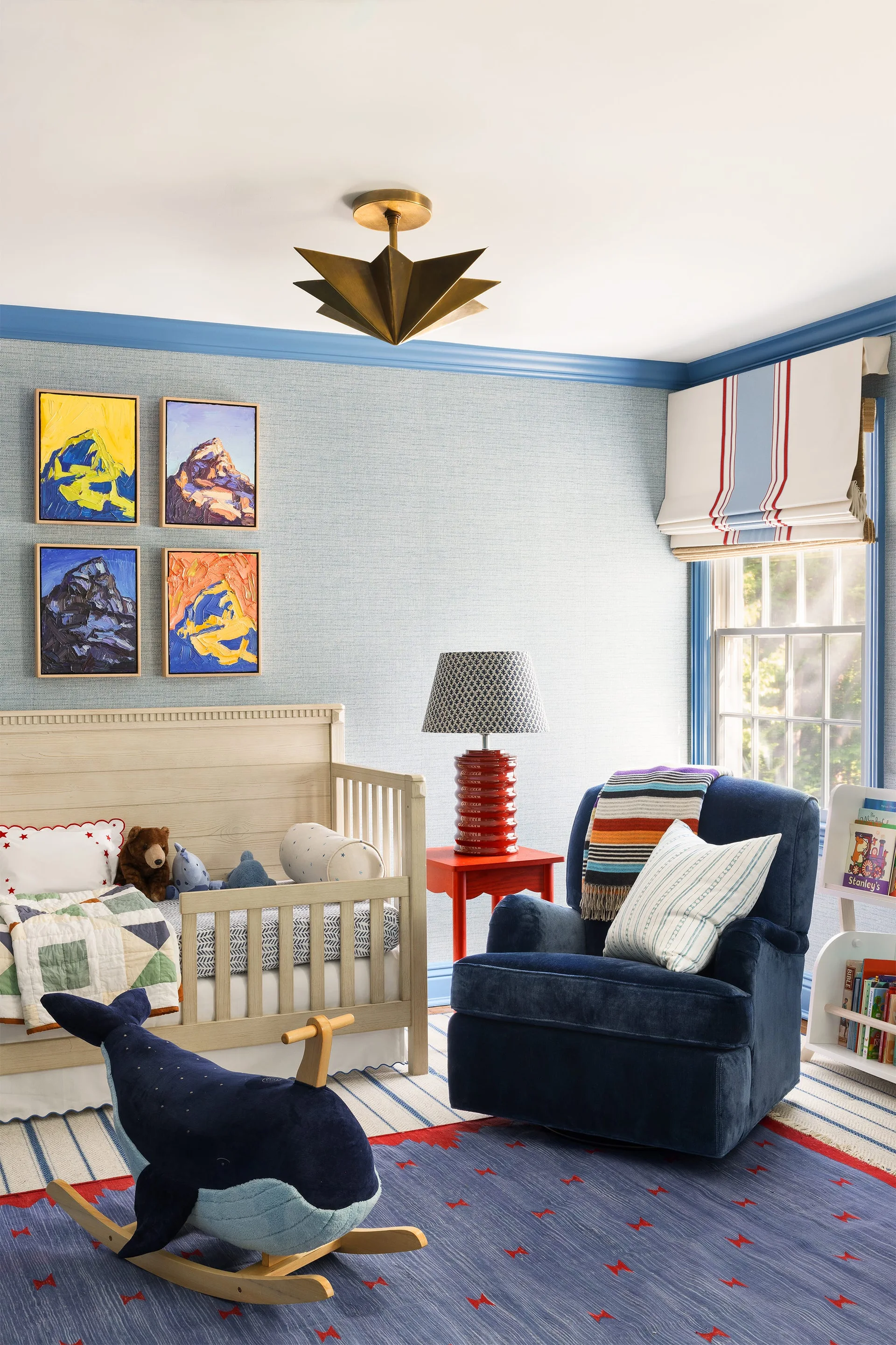 A nursery with blue textured wallpaper, a wooden crib, a navy blue swivel armchair, a red side table, and a starburst brass light fixture.