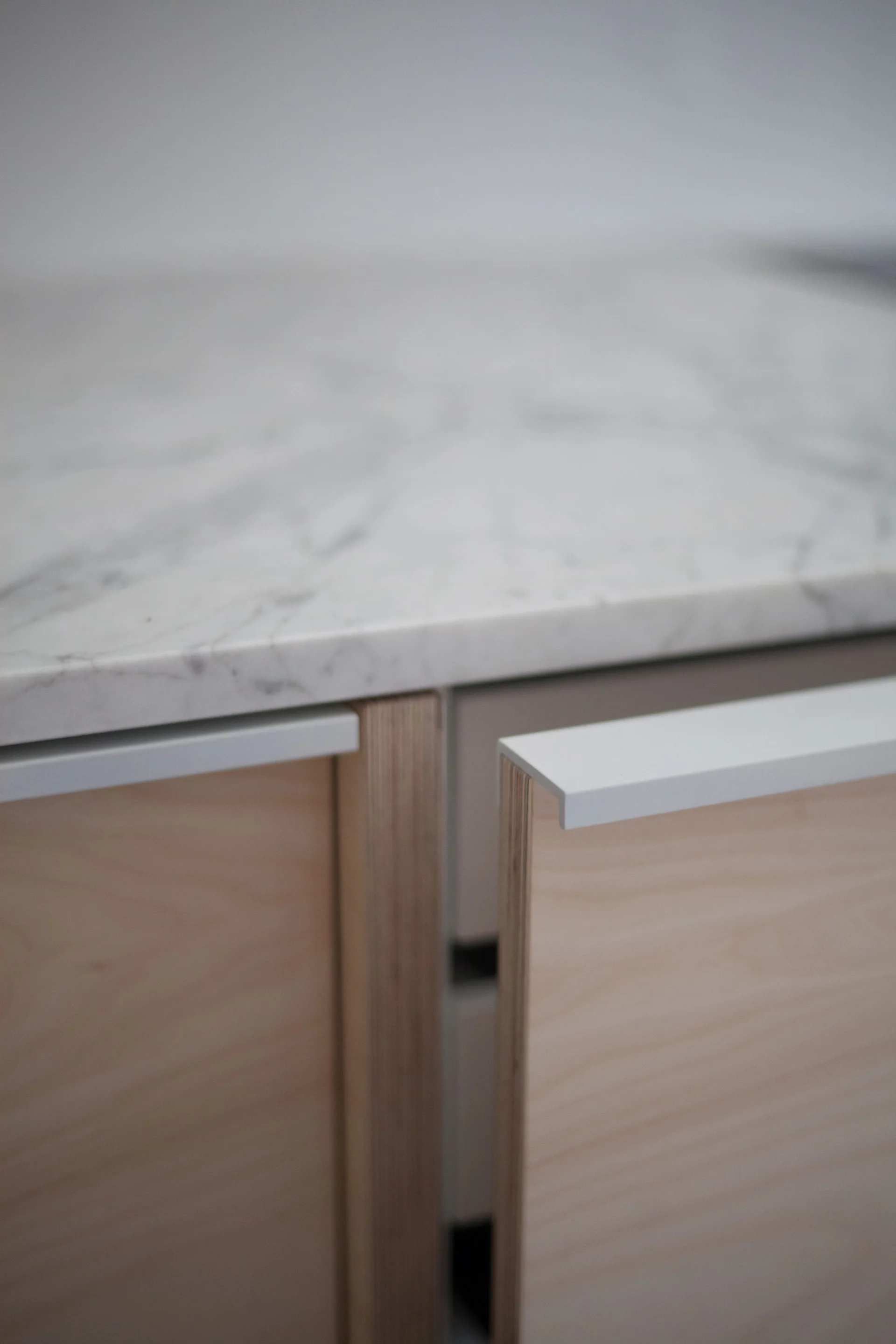 A close-up of a light wood plywood cabinet door slightly open under a veined white marble countertop with a white metal finger pull.