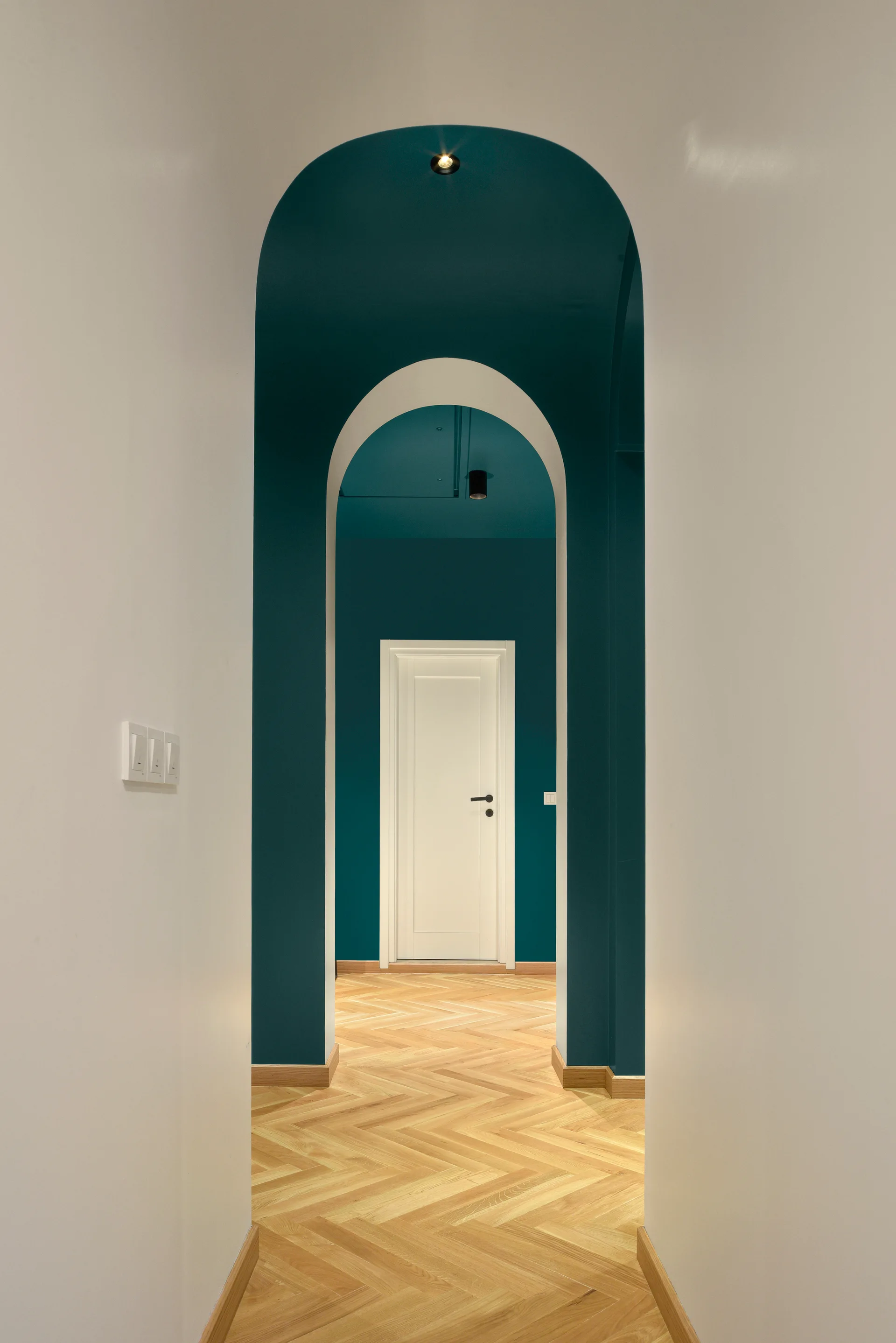 A narrow hallway featuring nested archways painted in deep teal, leading to a white interior door on herringbone patterned light wood floors.
