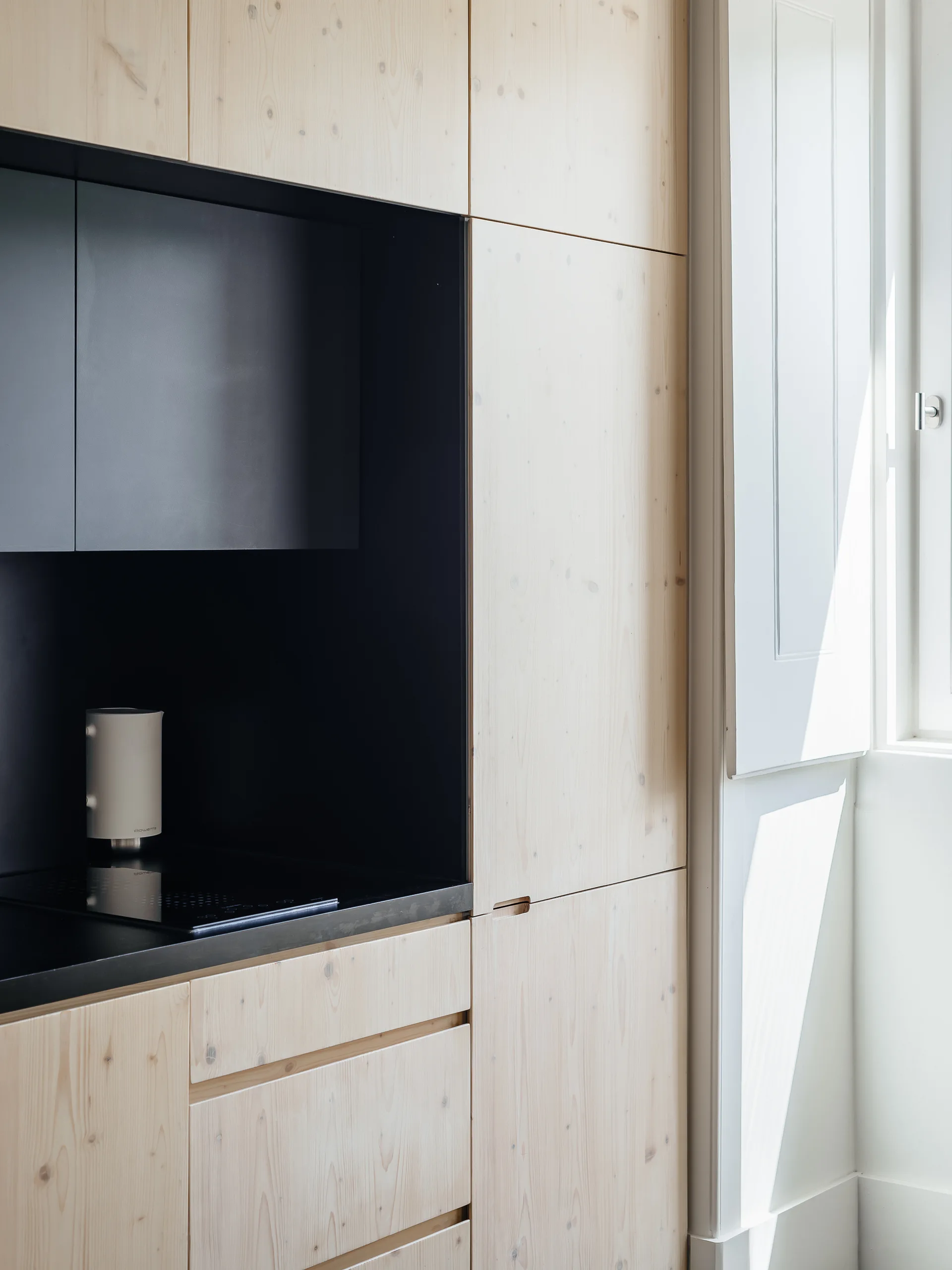 Flat-panel light wood cabinets surround a recessed matte black cooking niche with an induction cooktop next to a white paneled window shutter.