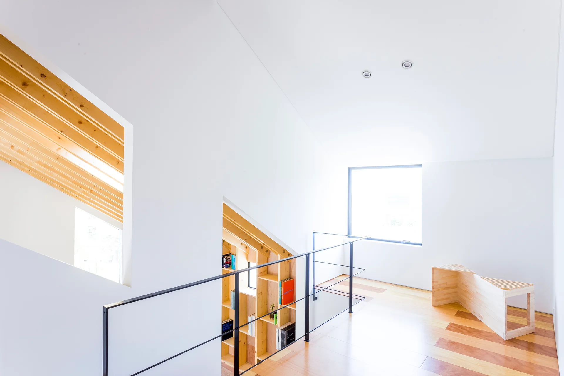 A minimalist white loft landing features warm wood floors, a black metal staircase railing, architectural cutouts, and a geometric wood bench.