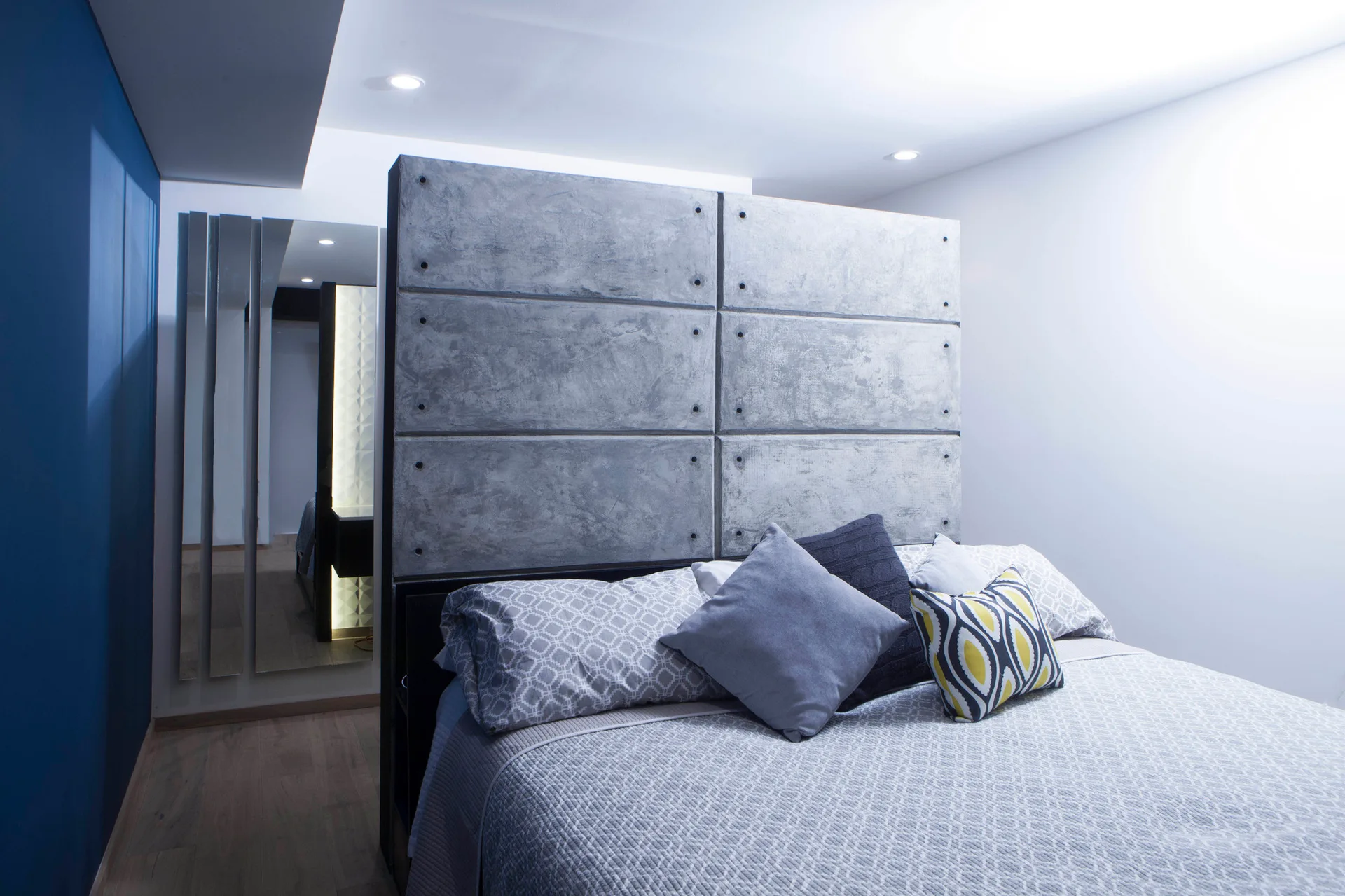 A modern bedroom features a large grey concrete panel headboard, textured bedding with geometric pillows, a deep blue wall, and vertical mirrors.