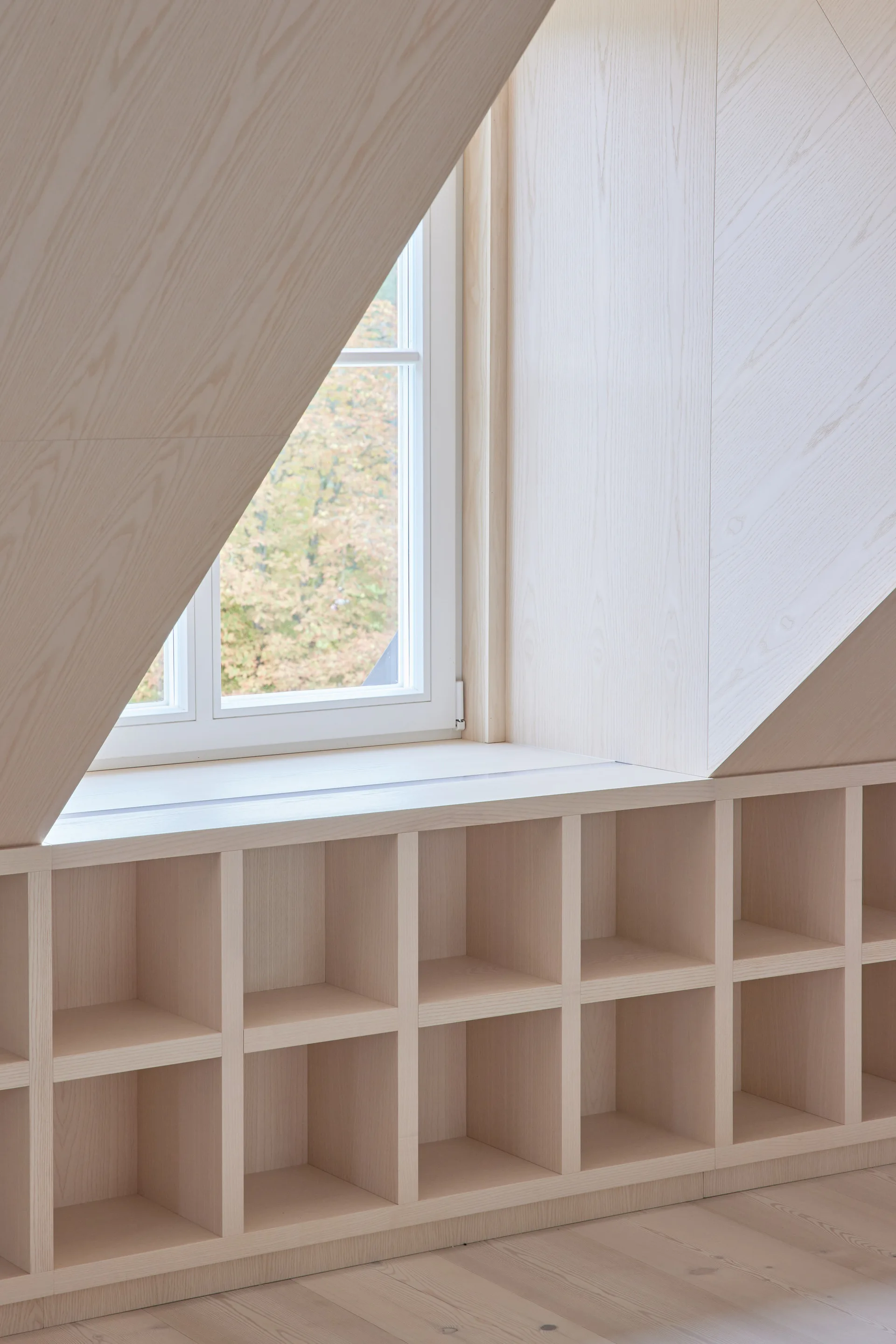 A minimalist attic room features a white-trimmed window flanked by sloped ceilings clad in light wood paneling, with built-in grid shelving below.