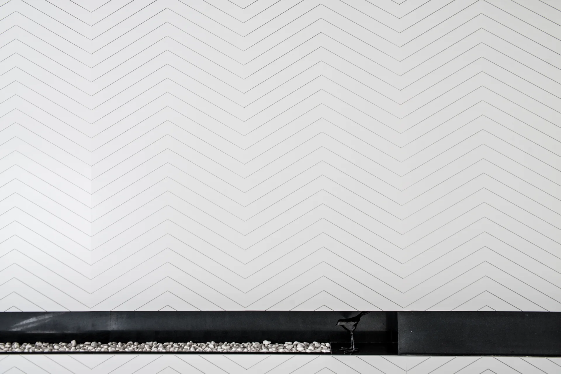 A white wall with a grey chevron pattern rises above a black recessed linear fireplace filled with small white river stones and a black bird sculpture.