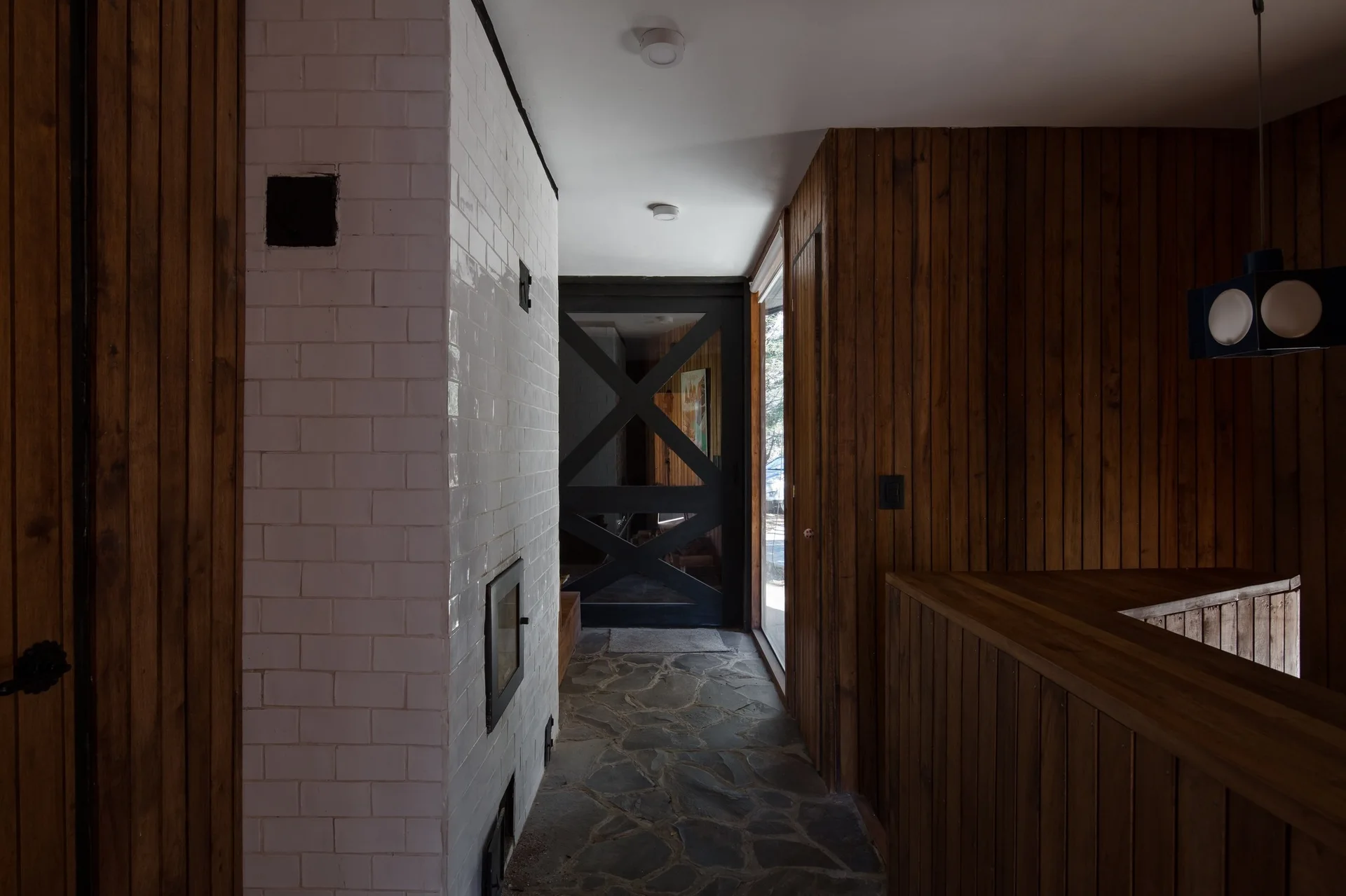 An entryway hallway with vertical wood paneling, grey stone flagstone flooring, a white tiled column, and a black front door with x-bracing.