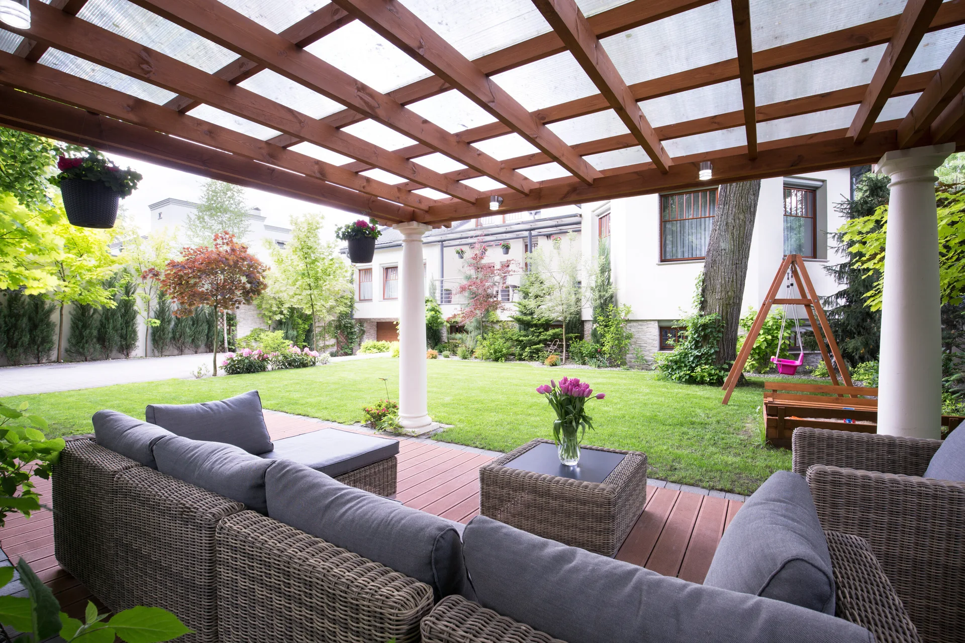 A covered wooden patio with a wicker sectional sofa overlooks a green grassy backyard with a swing set and garden.