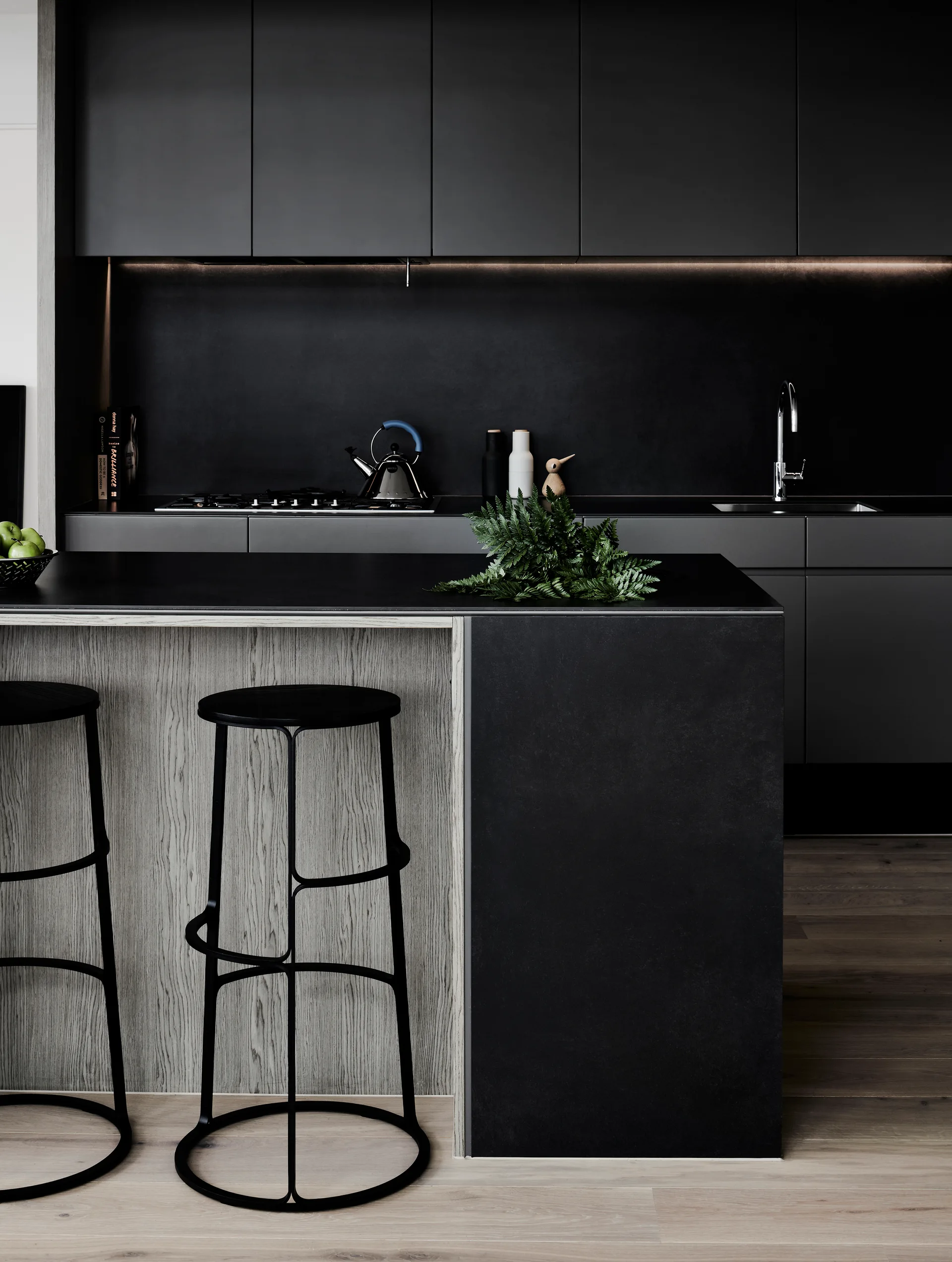 A modern kitchen with matte black upper cabinets, a matching black backsplash, and a kitchen island with a light wood grain panel and two black stools.