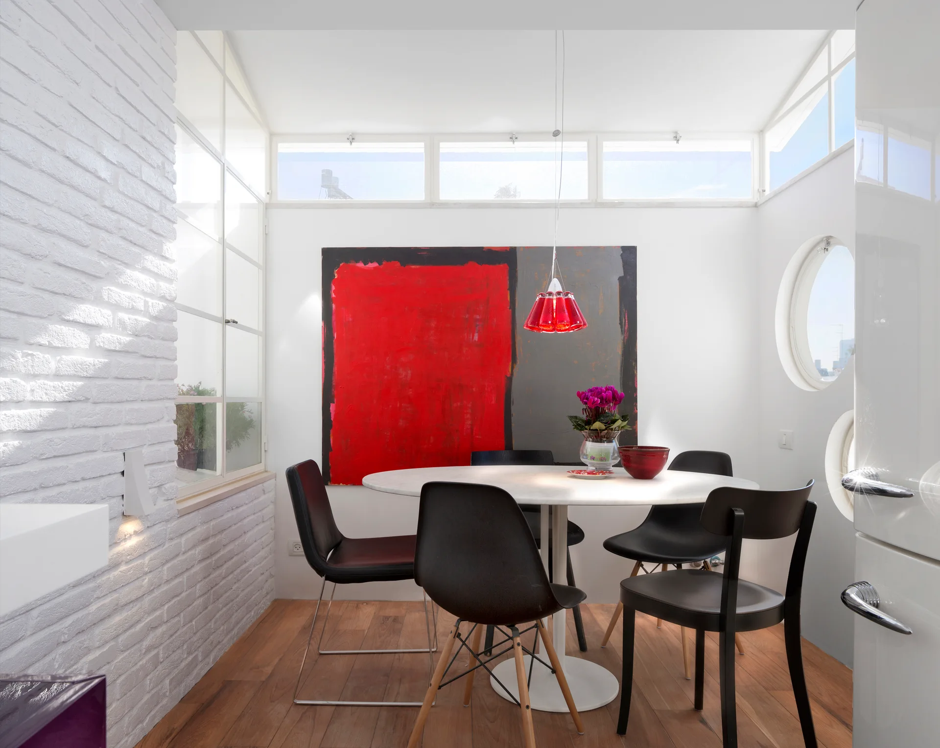 A round white table and black chairs sit on a wood floor below a red pendant light, backed by a large red abstract painting.