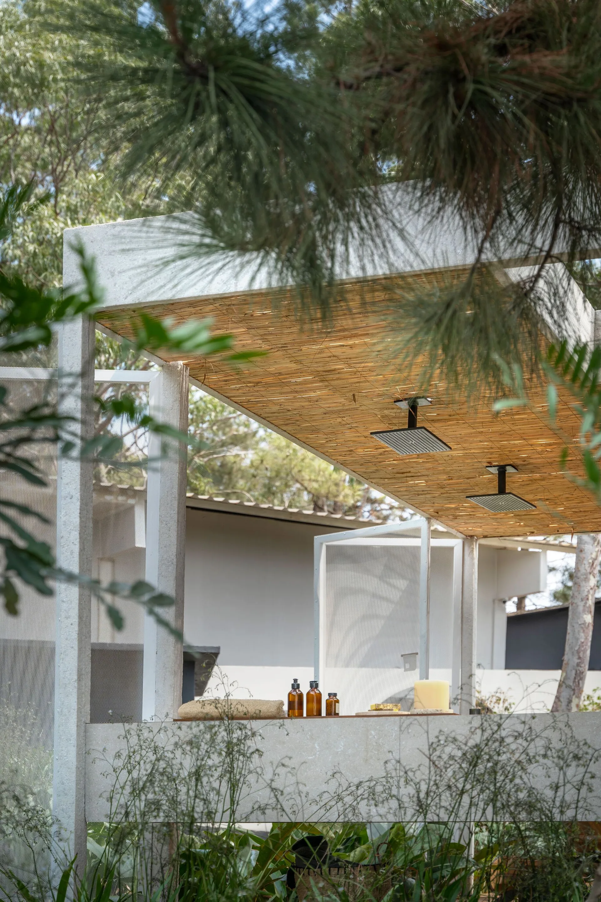 An outdoor shower area features a bamboo slatted ceiling with two rainfall showerheads, a concrete wall, and a ledge holding amber bottles and a candle.