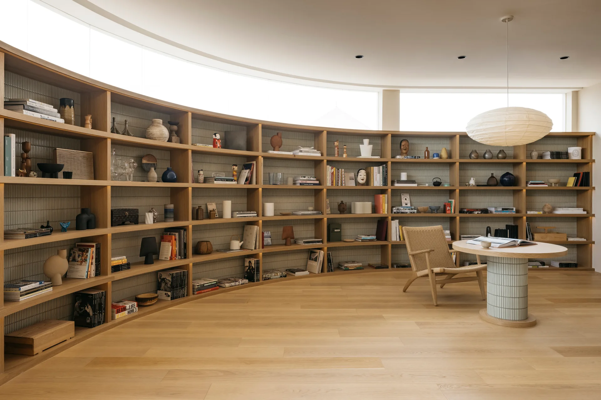 A large curved wooden bookshelf filled with books and decor in a room with a round tiled-base table and a woven armchair.