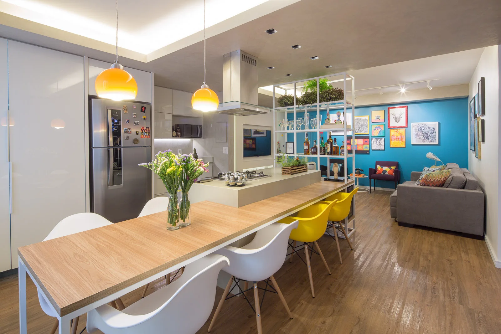 An open plan kitchen features a wood dining table attached to the island, yellow chairs, orange pendant lights, and a blue accent wall.