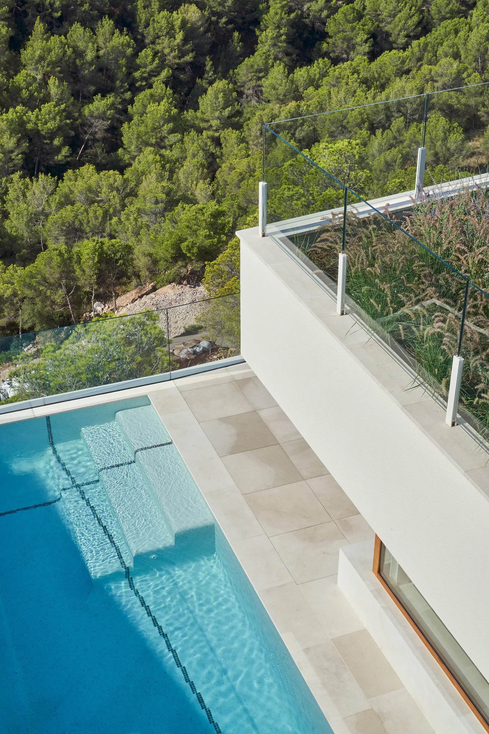 An aerial view of a modern outdoor swimming pool and concrete terrace with a glass railing overlooking a green hillside.
