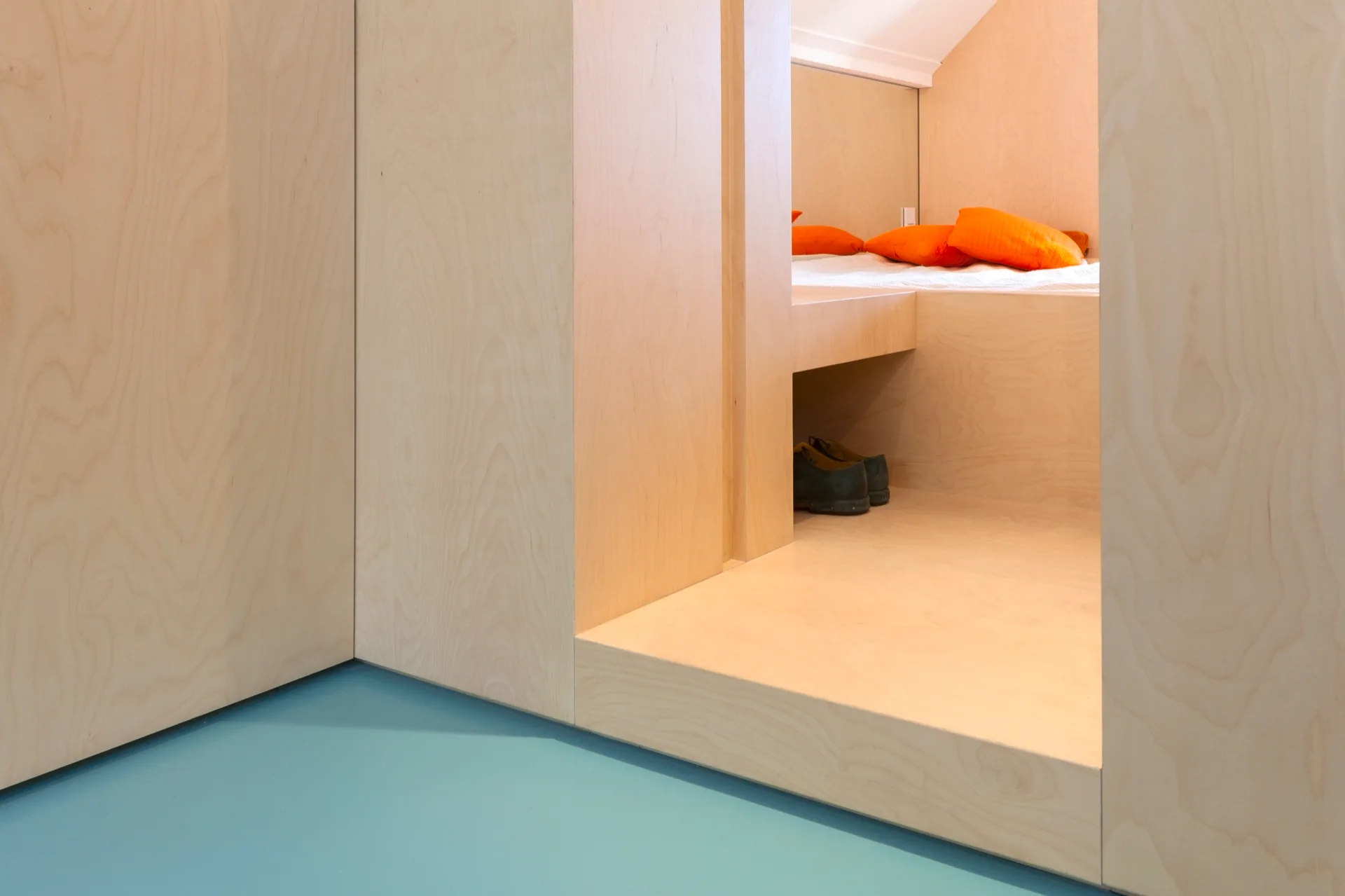 A minimalist bedroom interior features a built-in light plywood platform bed, bright orange pillows, an integrated cubby, and a smooth teal floor.