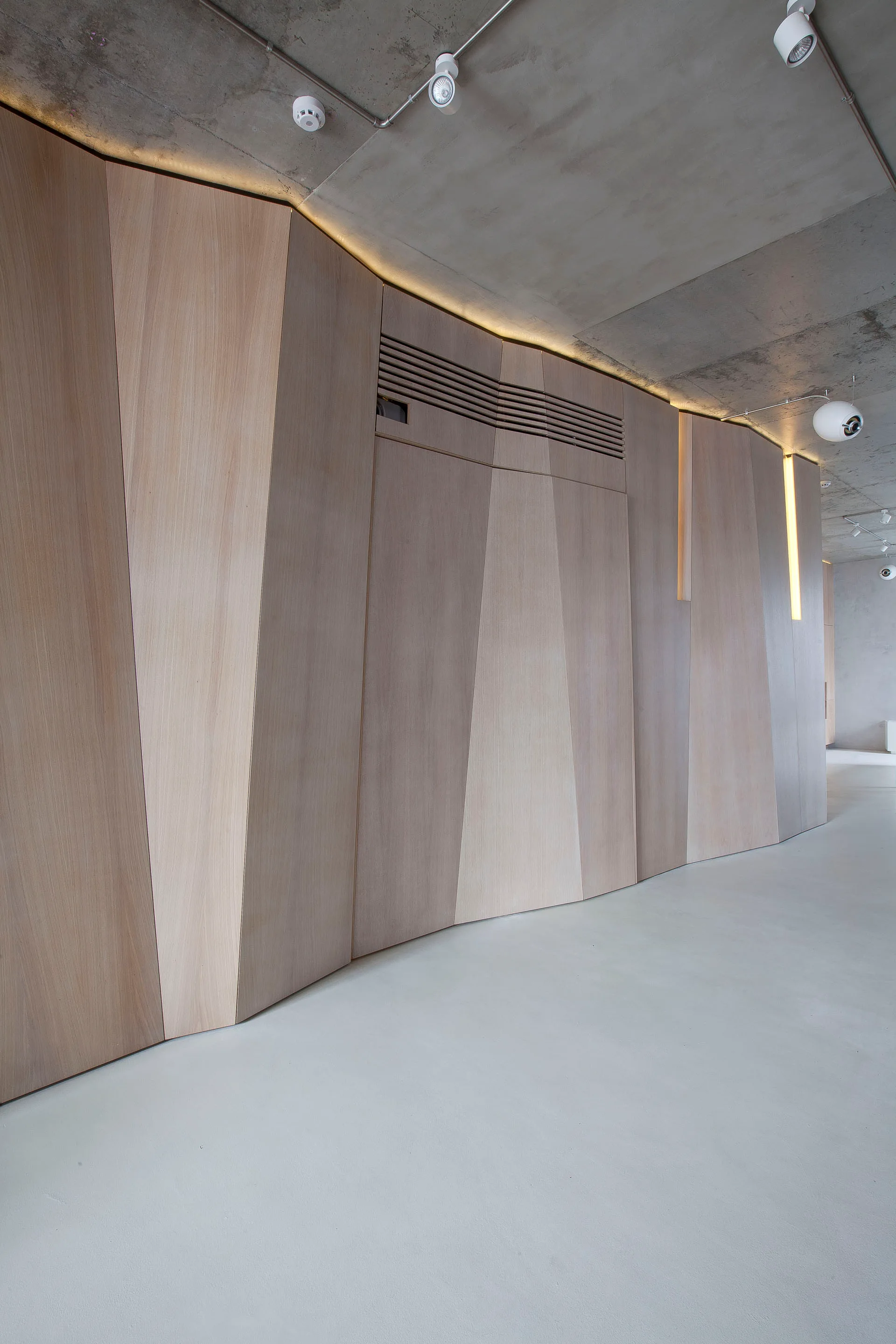 A sculptural wood-paneled wall with faceted geometric sections stands beneath a raw concrete ceiling in a minimalist hallway.