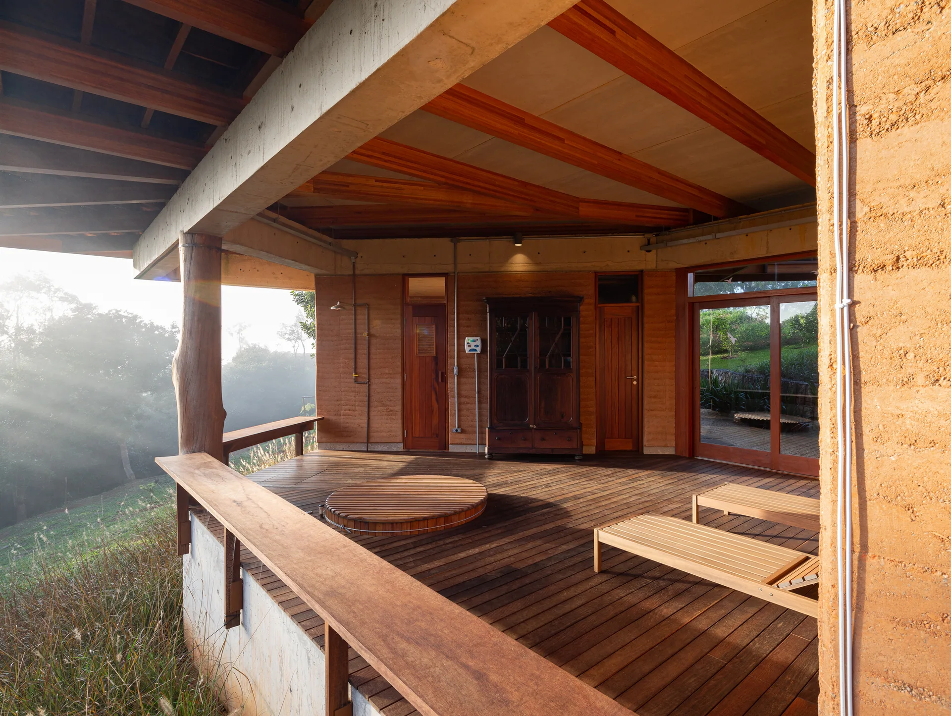 A covered wooden deck terrace features a circular in-floor hot tub, lounge chairs, and textured terracotta-colored rammed earth walls.