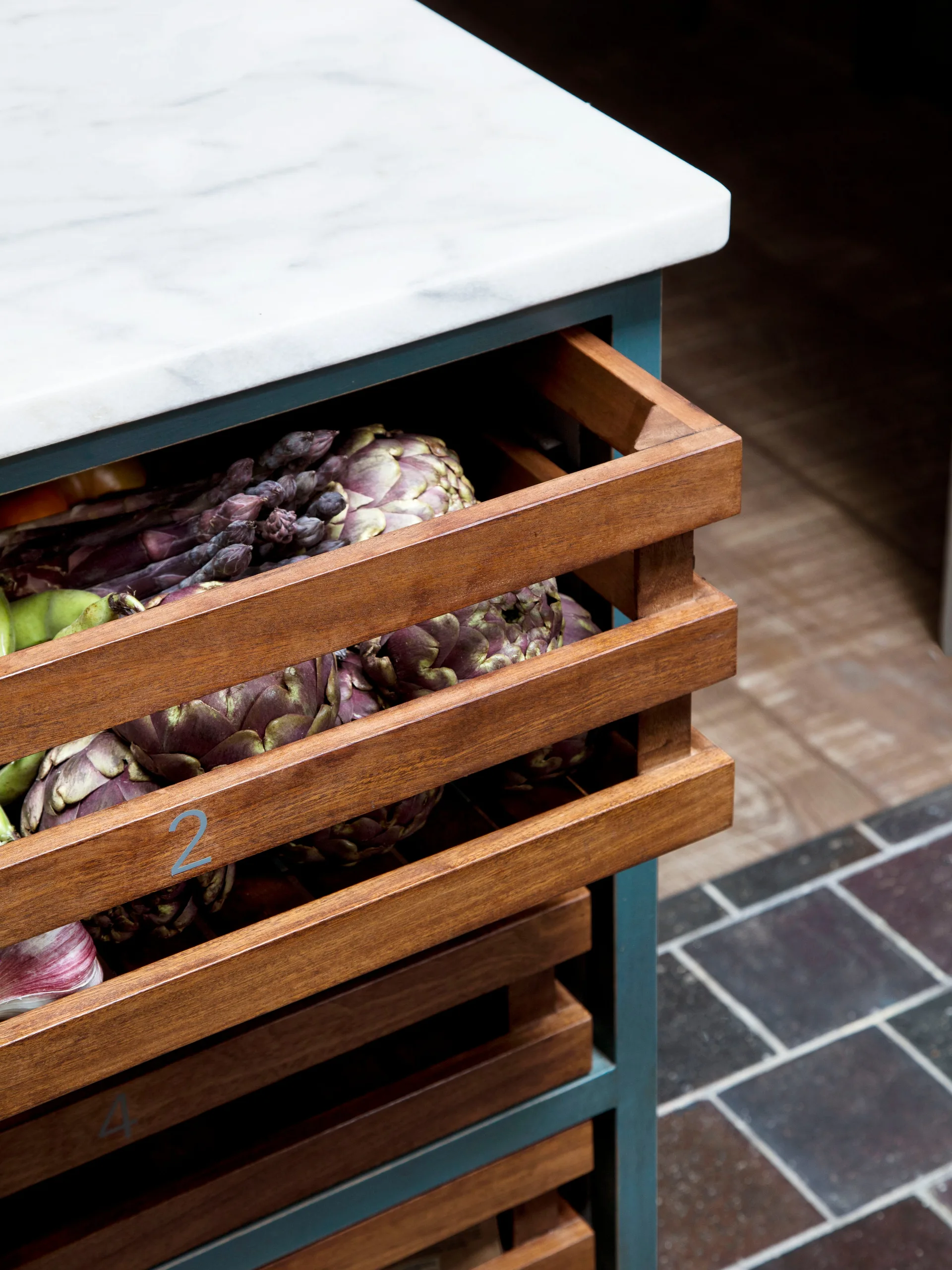 A close-up of a teal kitchen island featuring pull-out slatted wooden crates filled with artichokes beneath a white marble countertop.