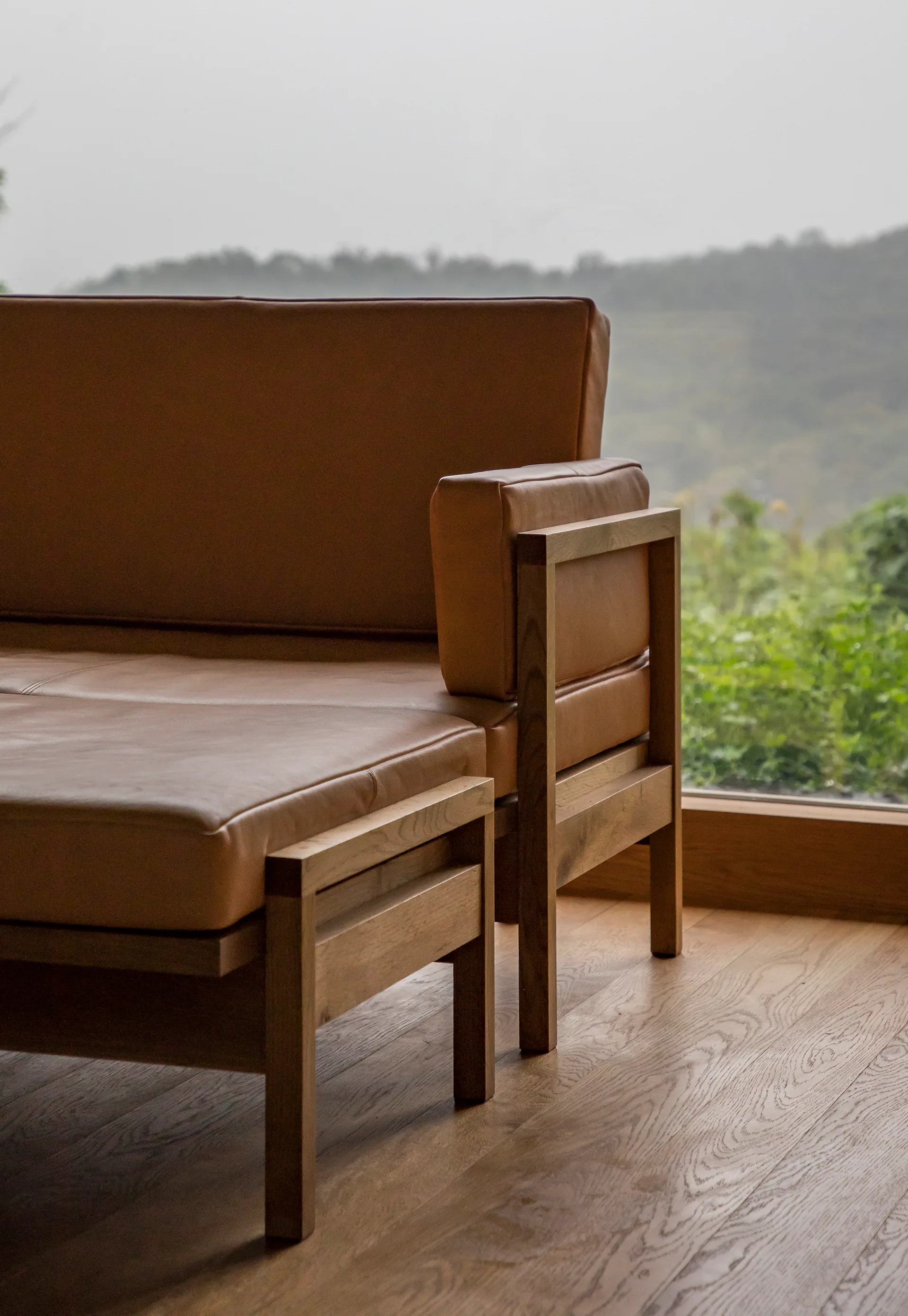 A tan leather sofa with a light oak wood frame stands on hardwood floors in front of a window overlooking a green landscape.