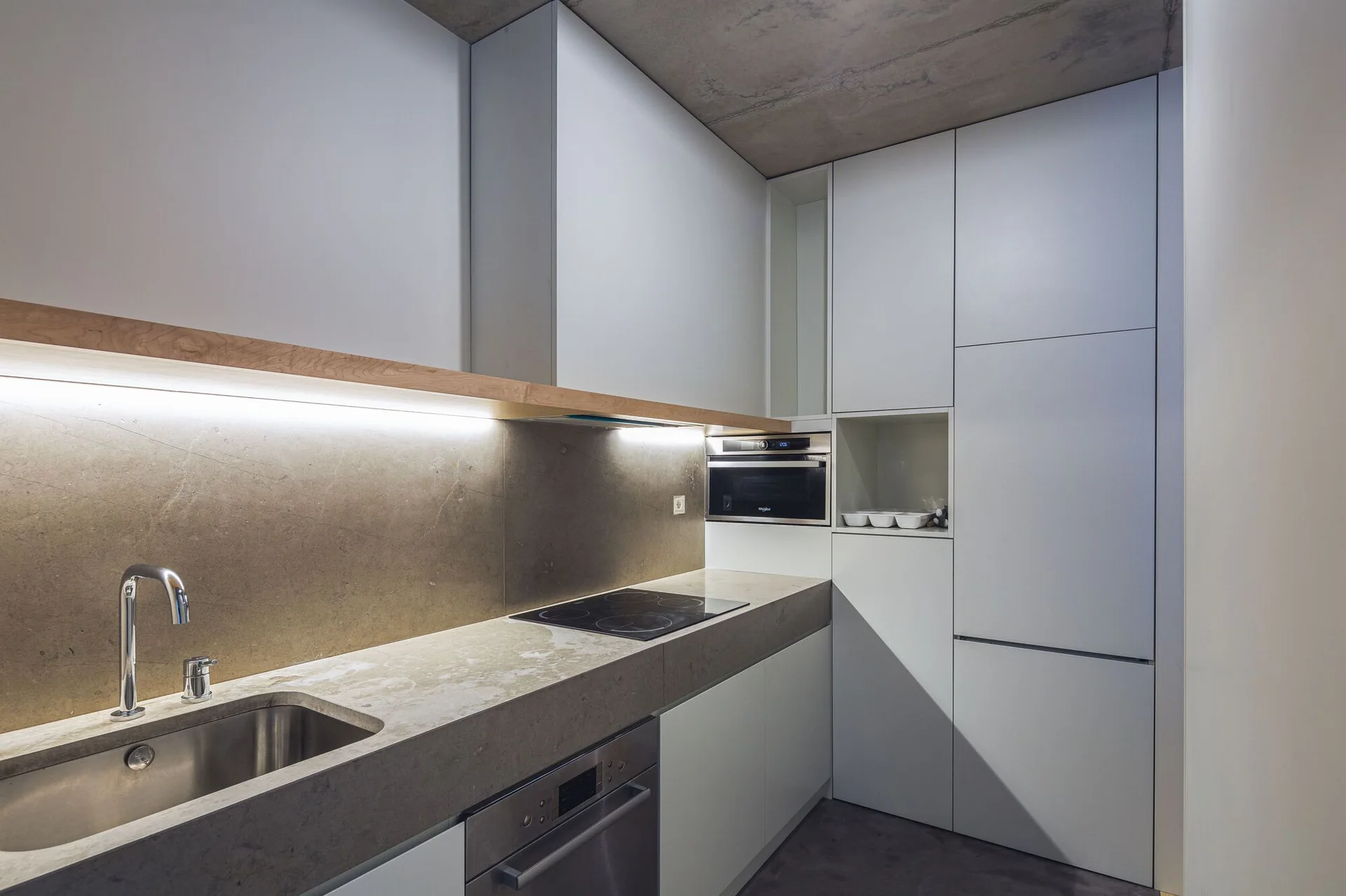 A modern minimalist kitchen featuring white flat-panel cabinets, a thick stone-like countertop, and an exposed raw concrete ceiling.