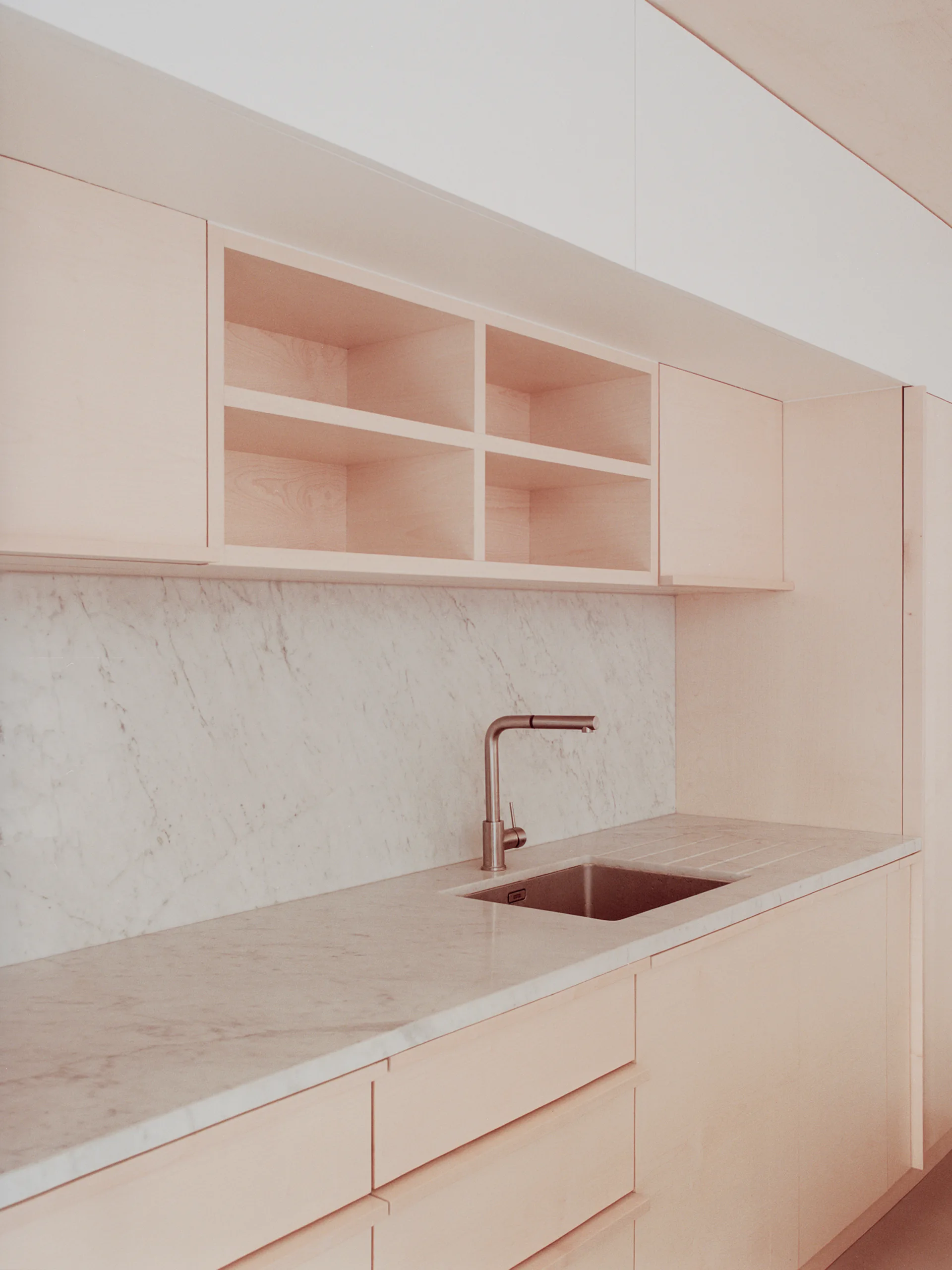 A minimalist kitchen features light-toned wood cabinetry with open shelving, a white marble backsplash, and an undermount sink with a metallic faucet.