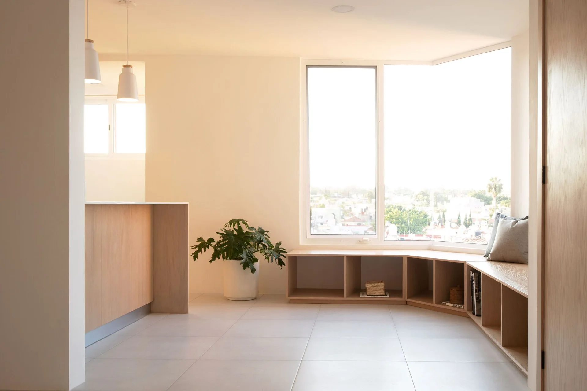 A minimalist room with a light wood built-in bench and shelving unit wrapping beneath an angled bay window, alongside large tiled flooring.