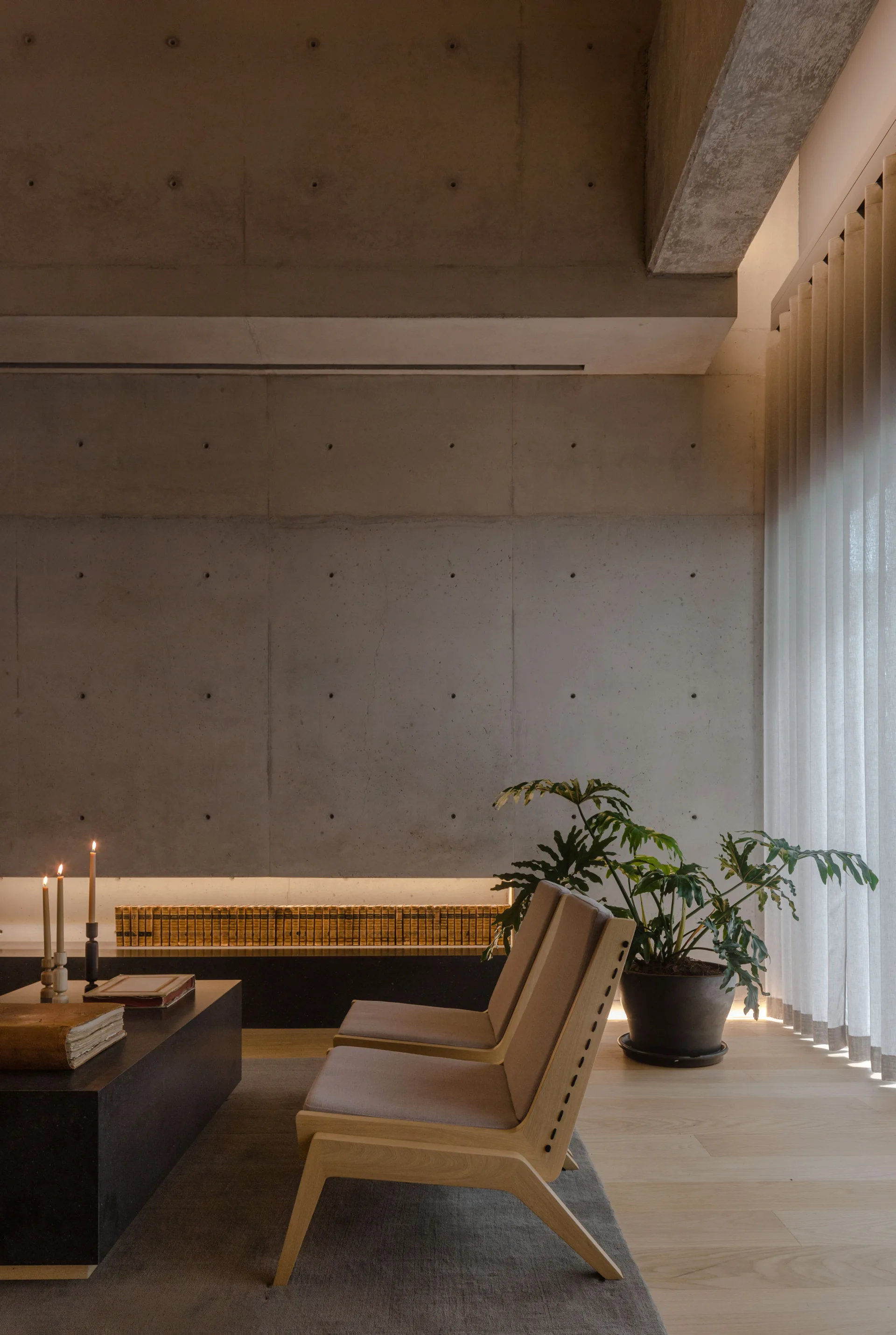 Two low wood lounge chairs sit in a minimalist living room with an exposed concrete wall, a low black coffee table, and sheer curtains.