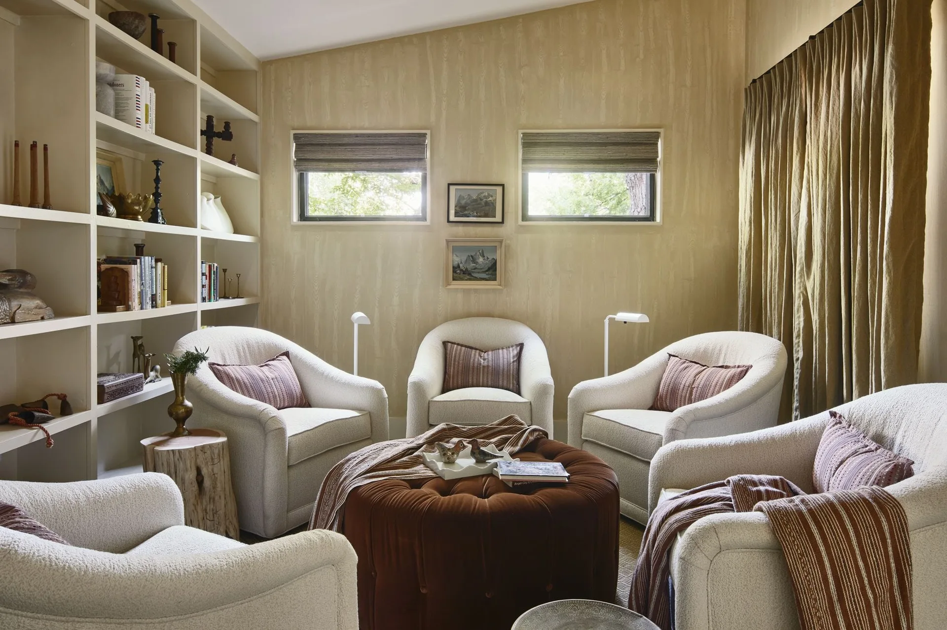 Five cream-colored boucle armchairs are arranged in a tight circle around a large brown tufted velvet ottoman in a cozy wood-paneled library.