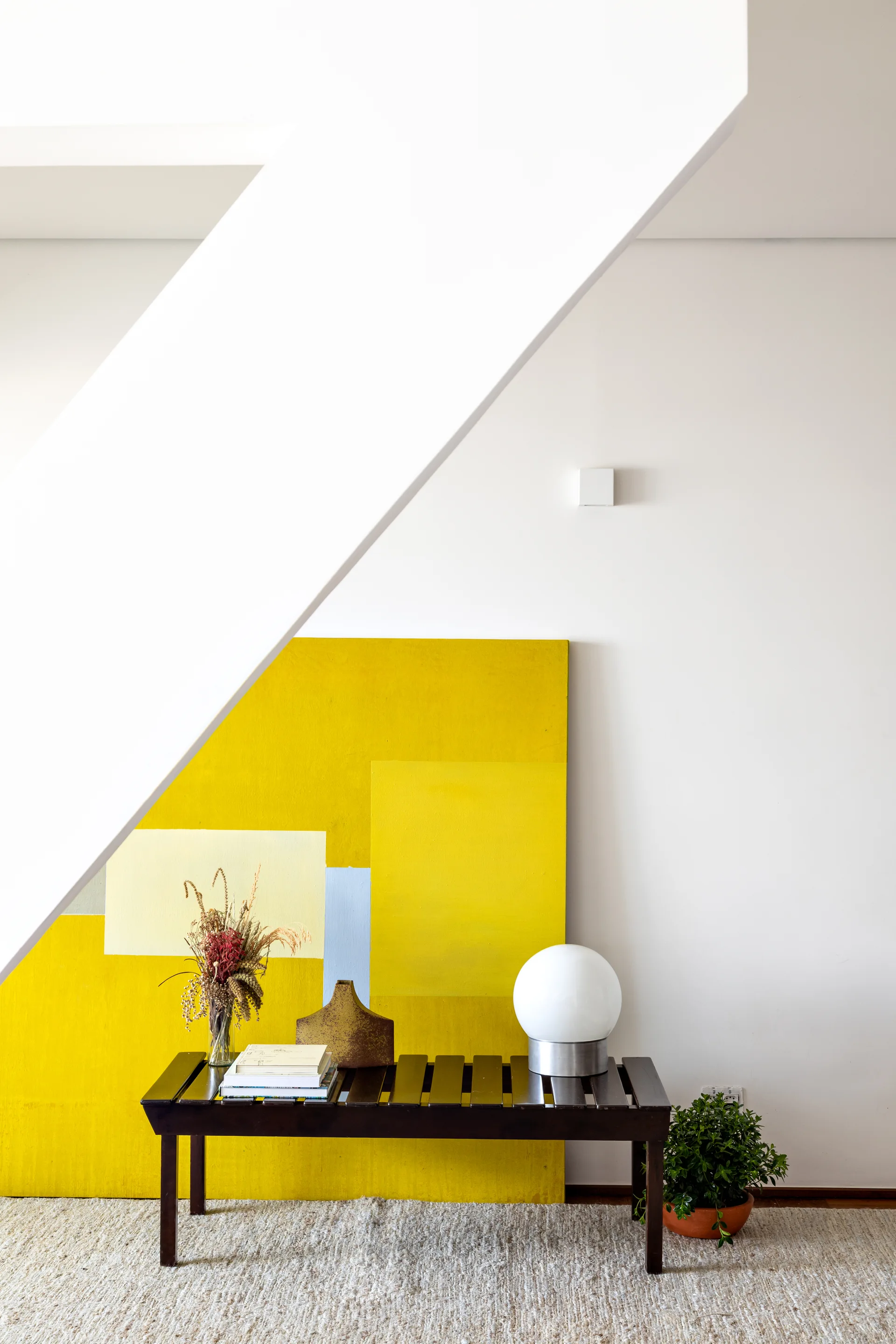 A dark wood slatted bench sits against a white wall under an angled staircase beam, displaying a globe lamp and a large yellow abstract painting.